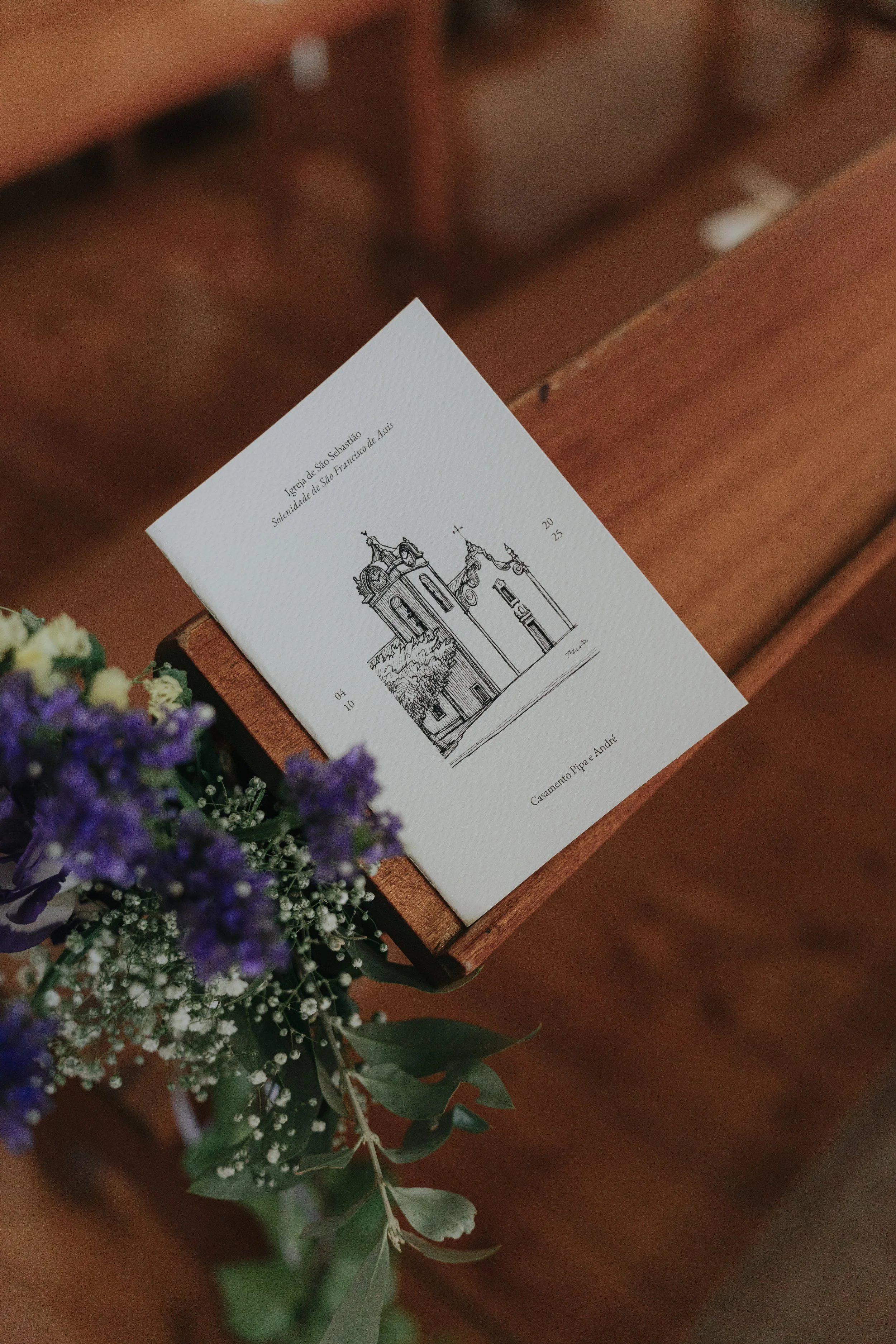 An invitation card with a line drawing of a church tower, placed on a wooden surface. There are purple and white flowers in the foreground.