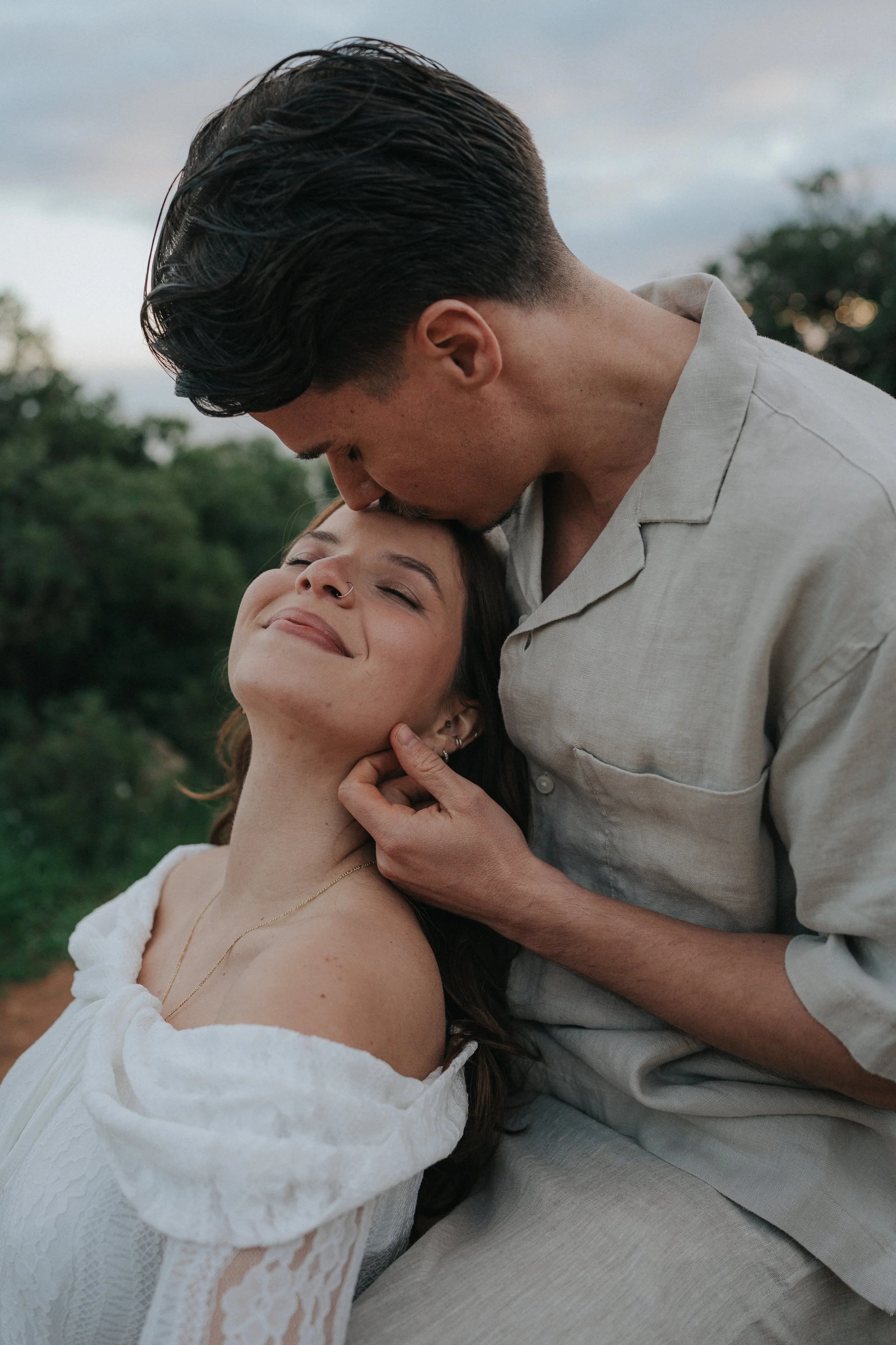 A man and woman share an intimate moment outdoors, with the man gently kissing the woman's forehead while she smiles with closed eyes. The woman has a nose ring and wears a white lace dress, and the man has dark, slicked-back hair and wears a light-c