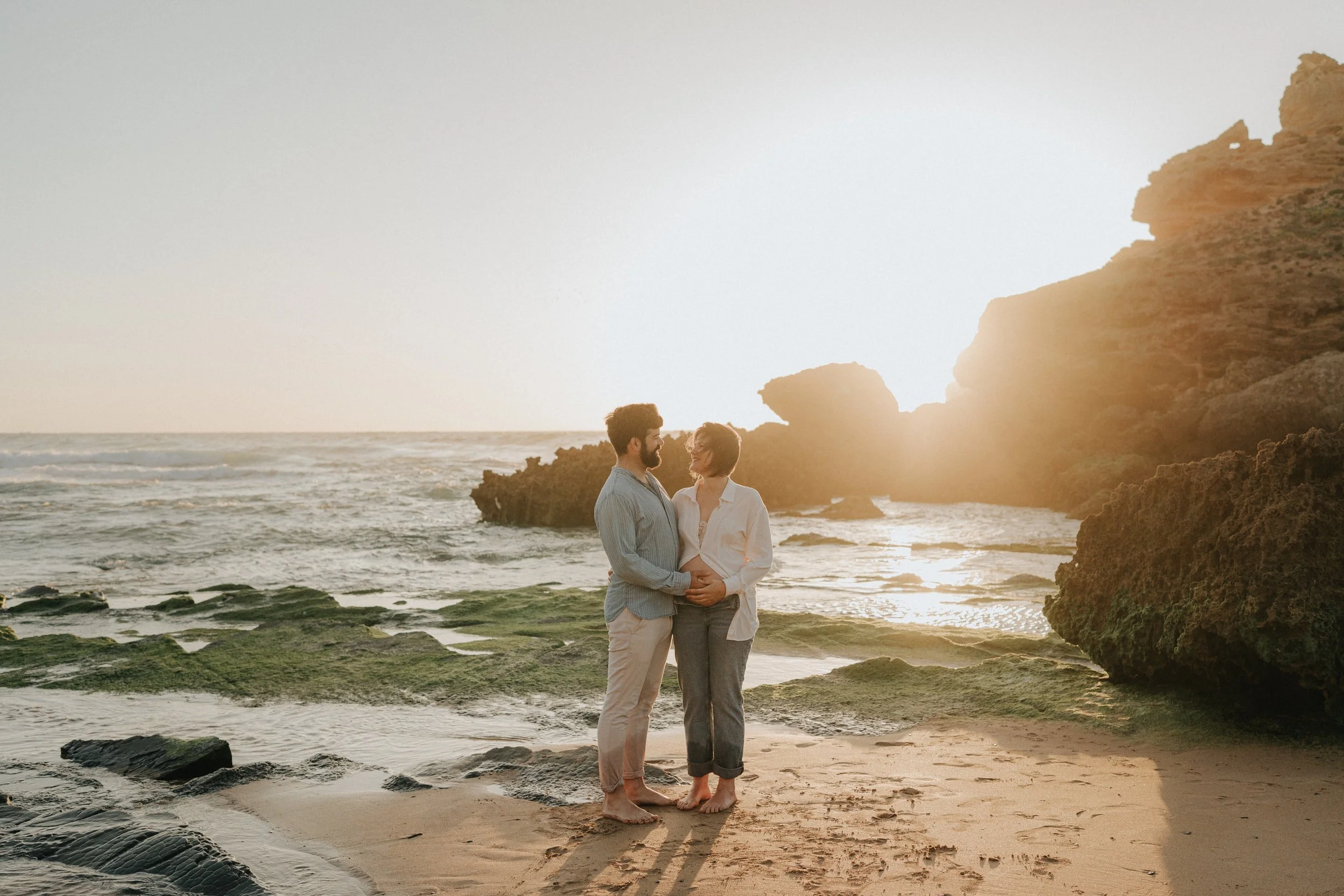 A couple standing on a beach at sunset, holding hands and gazing at each other, with rocky cliffs in the background and the ocean waves around their feet.