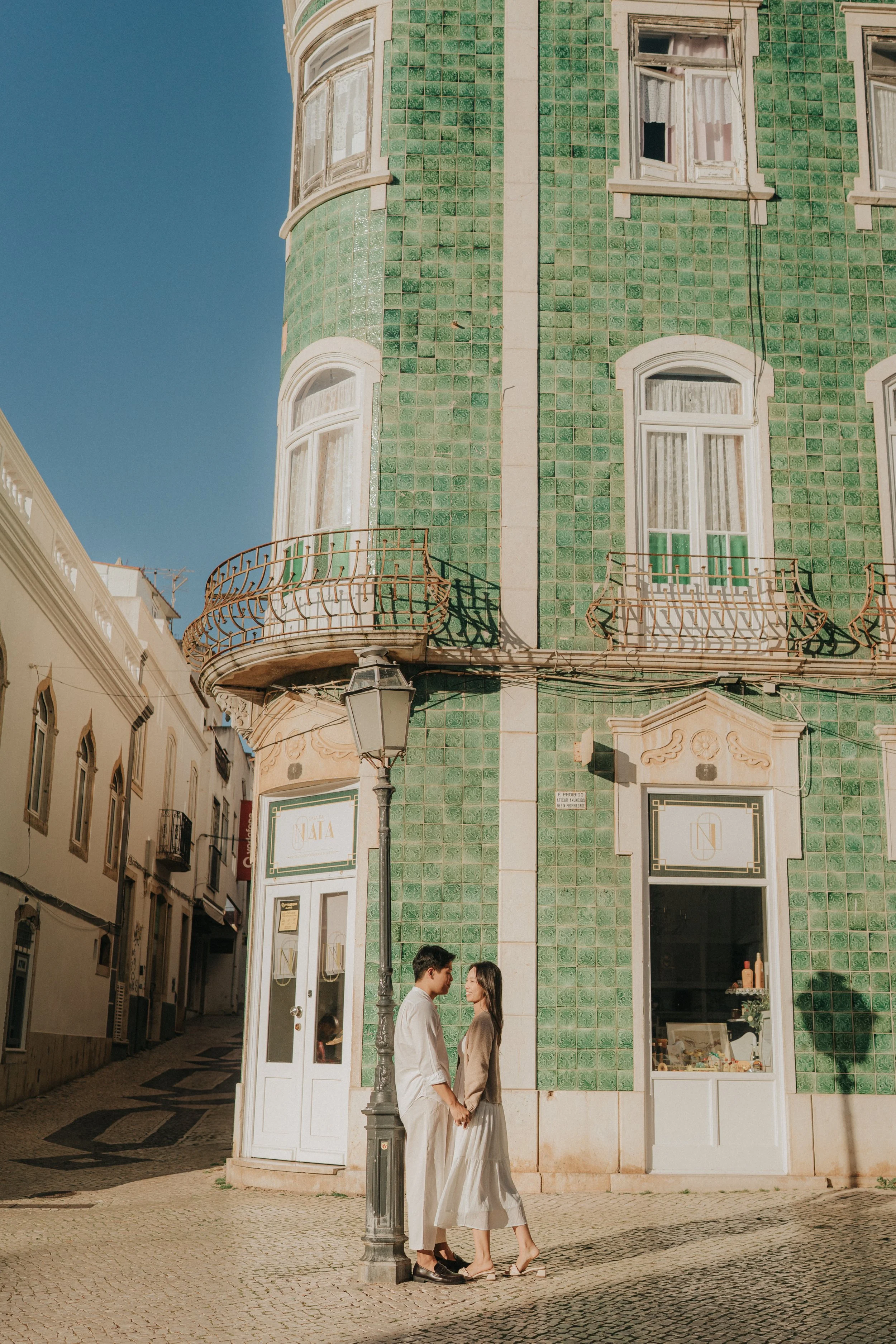 A couple is standing close together, holding hands, on a cobblestone street in front of a vintage building with a green tiled facade, white trim, and large windows. The man is wearing a white shirt and beige pants, and the woman is wearing a beige ja