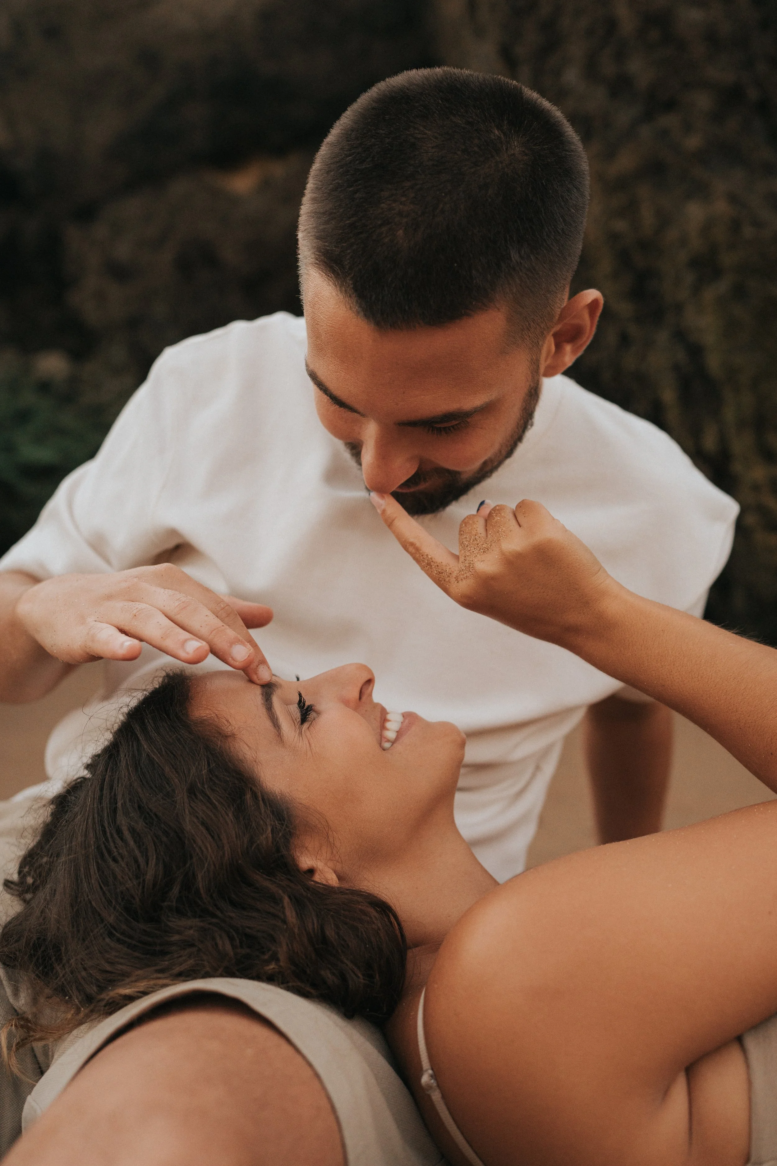 A man and a woman are close together, smiling as the man gently touches the woman's forehead with his finger. They seem to be in a warm, intimate moment.