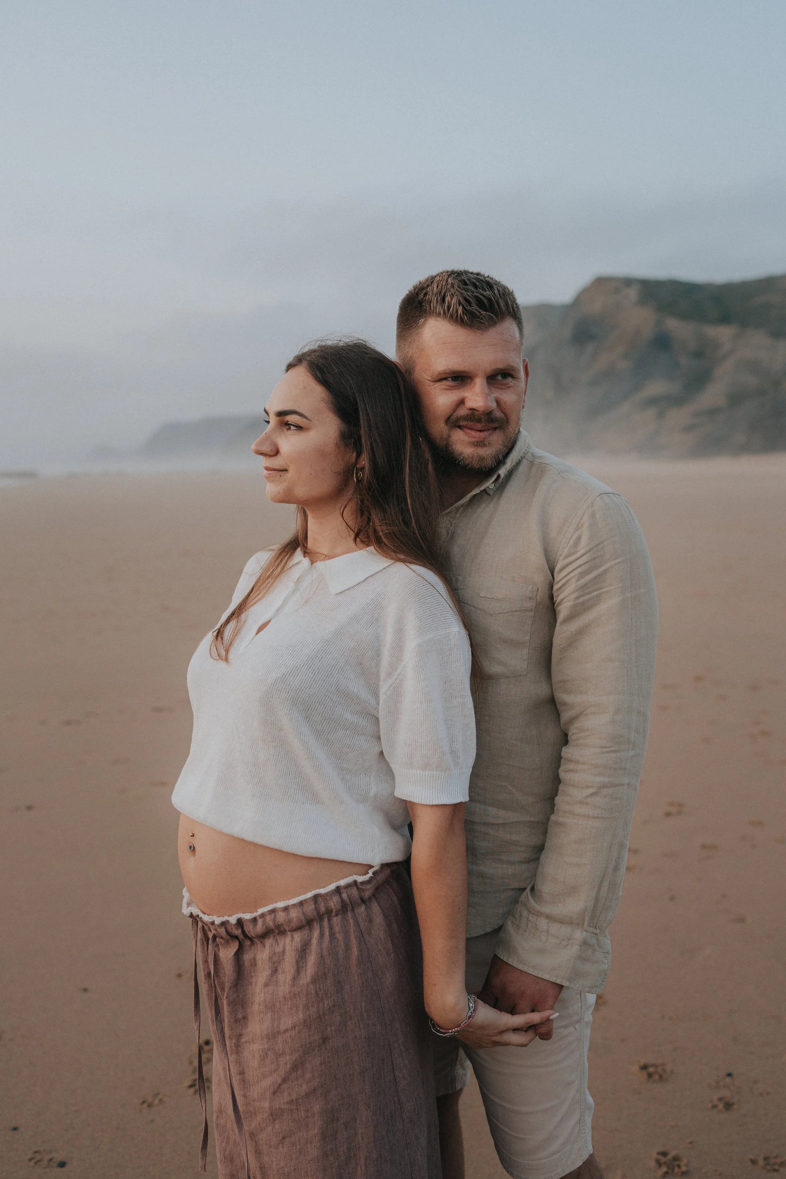 A man and a woman standing on a beach, facing opposite directions with the woman looking to the left and the man looking to the right, holding hands, with cliffs in the background.
