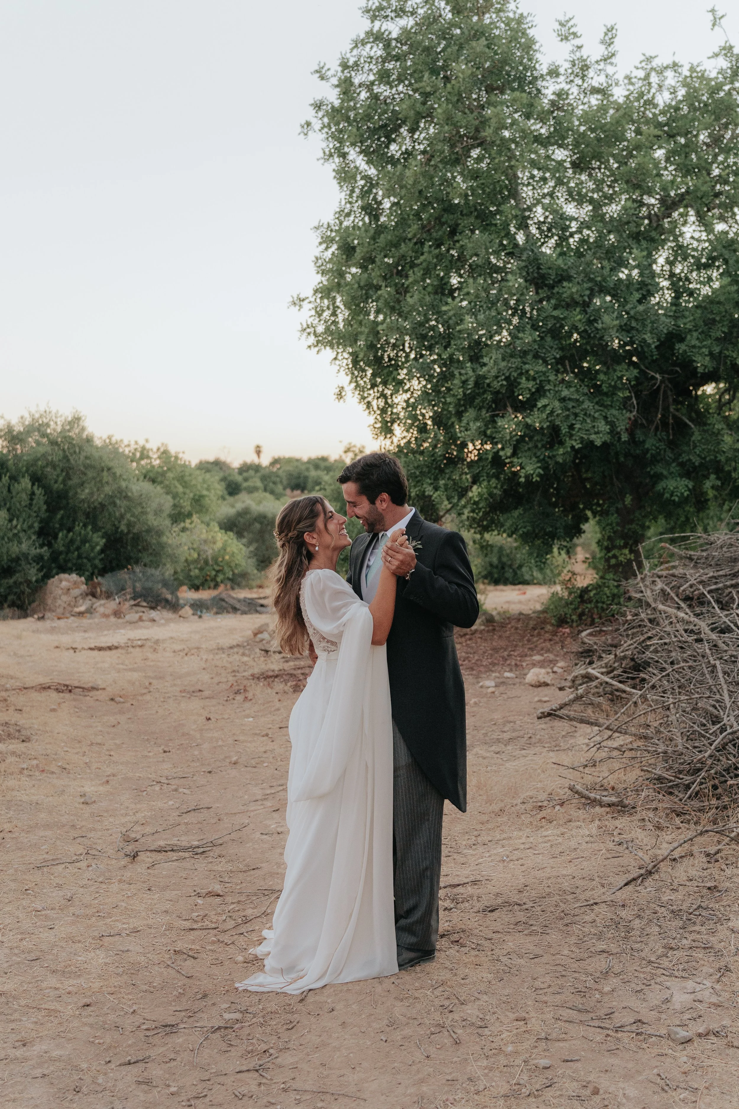 A bride and groom dance outdoors on dry, rocky ground near a large tree at dusk, with a clear sky and trees in the background.
