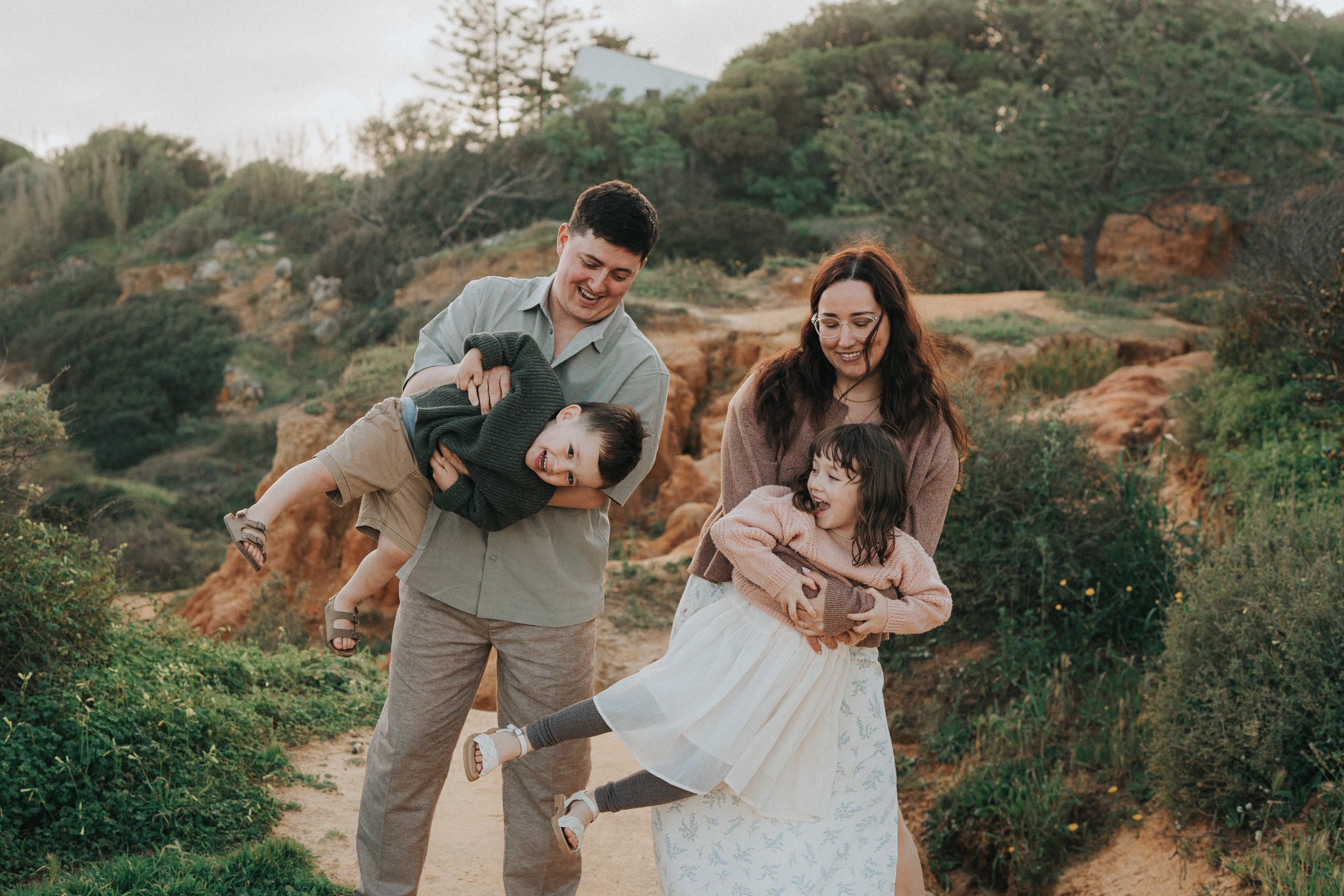 A family of four, including a man, woman, and two young children, smiling and playing outdoors on a dirt trail surrounded by greenery and shrubs.