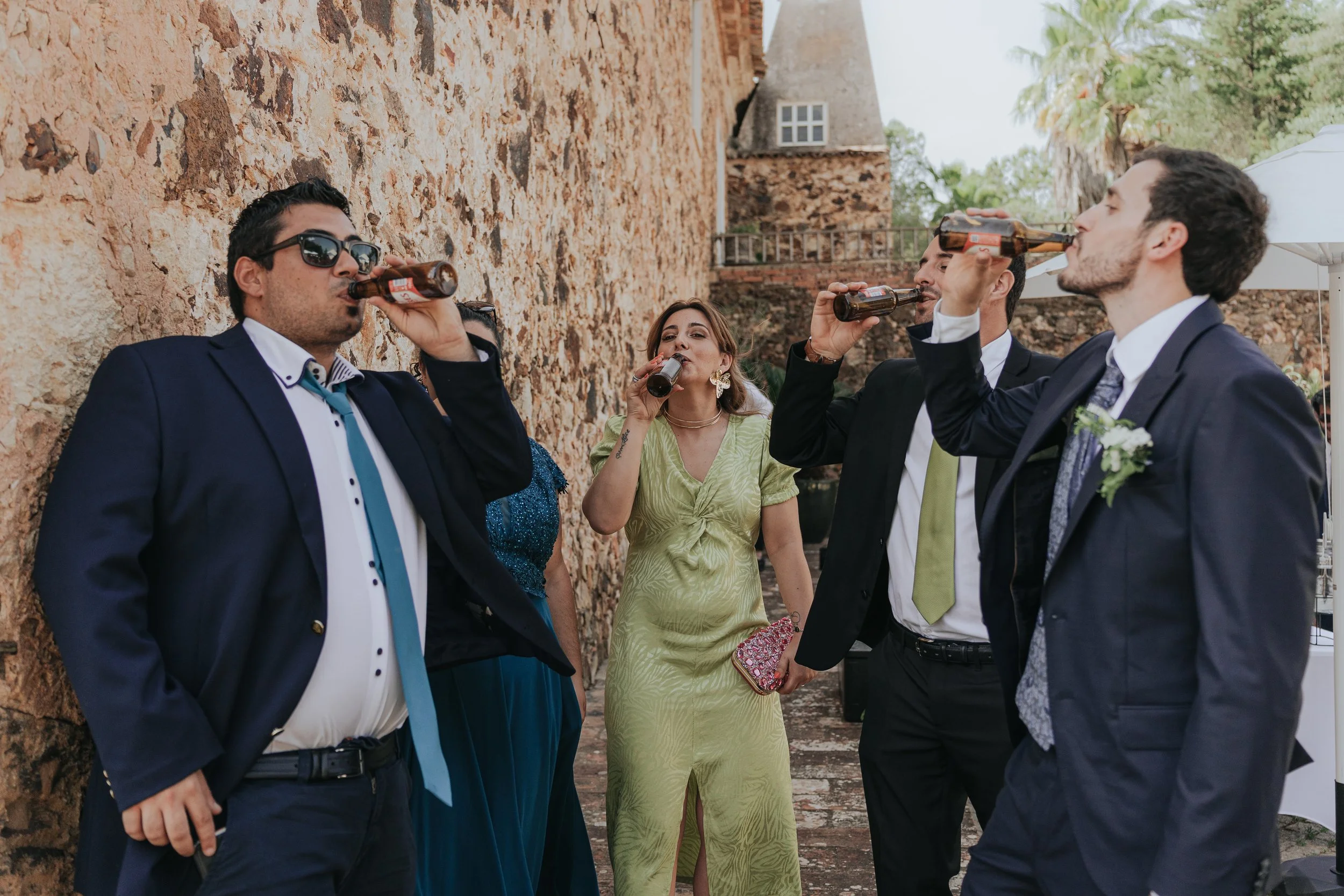 Group of young adults dressed in formal attire, enjoying drinks outdoors next to a brick wall with trees in the background.
