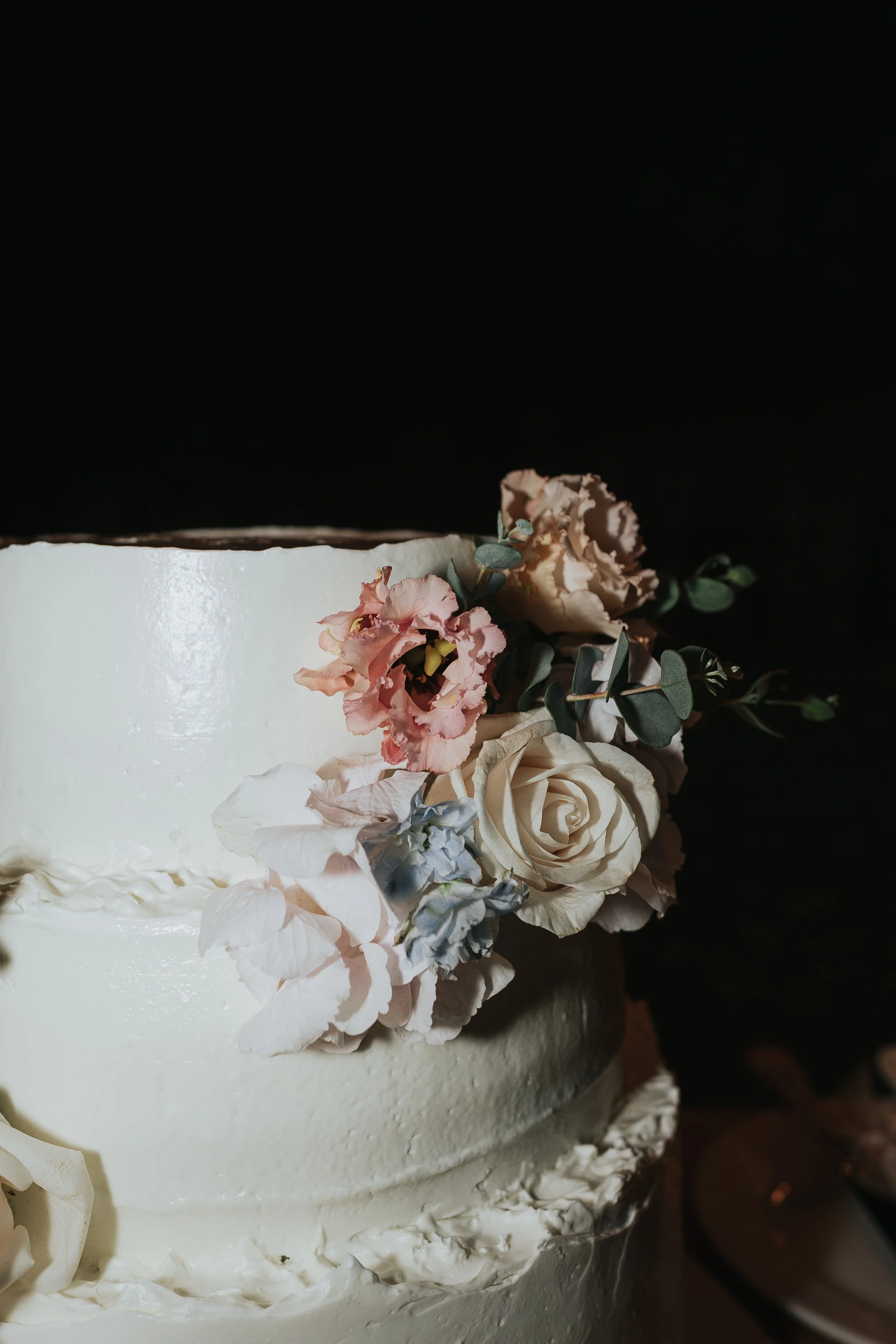 Close-up of a white cake decorated with a variety of pastel-colored flowers and green foliage against a black background.