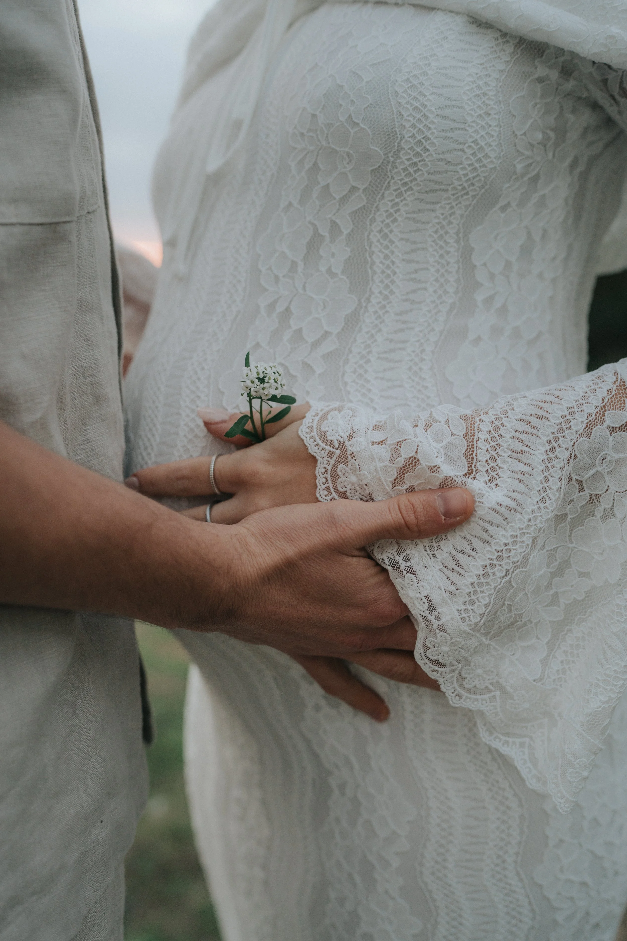 Close-up of a bride and groom holding hands, with the bride holding a small white flower and greenery. The bride is wearing a lace wedding dress, and the groom is in a light-colored suit. Their hands are gently clasped, capturing a romantic moment.