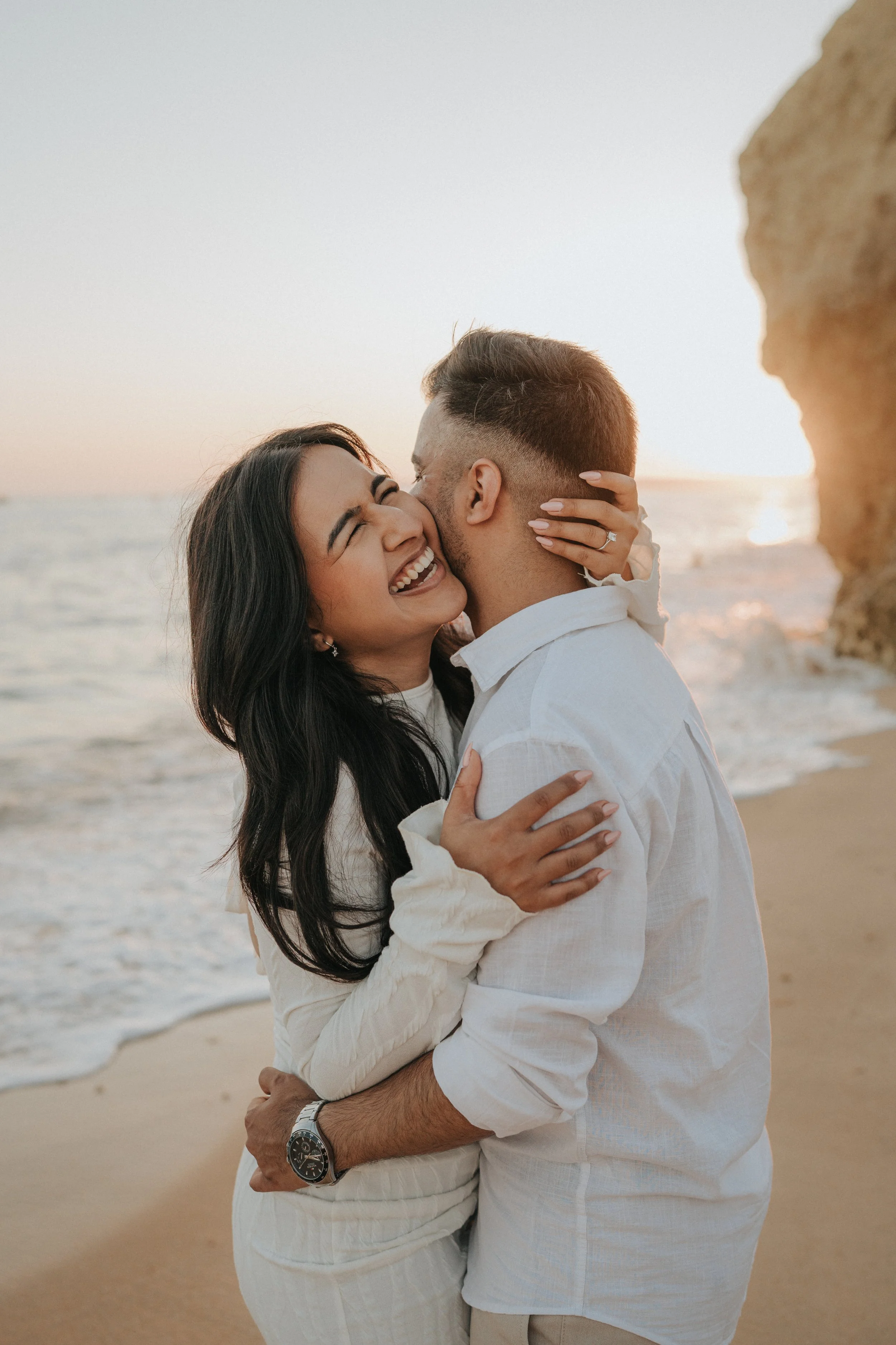 A couple hugging and kissing on the beach during sunset, with the ocean and sky in the background and a rocky formation on the right.