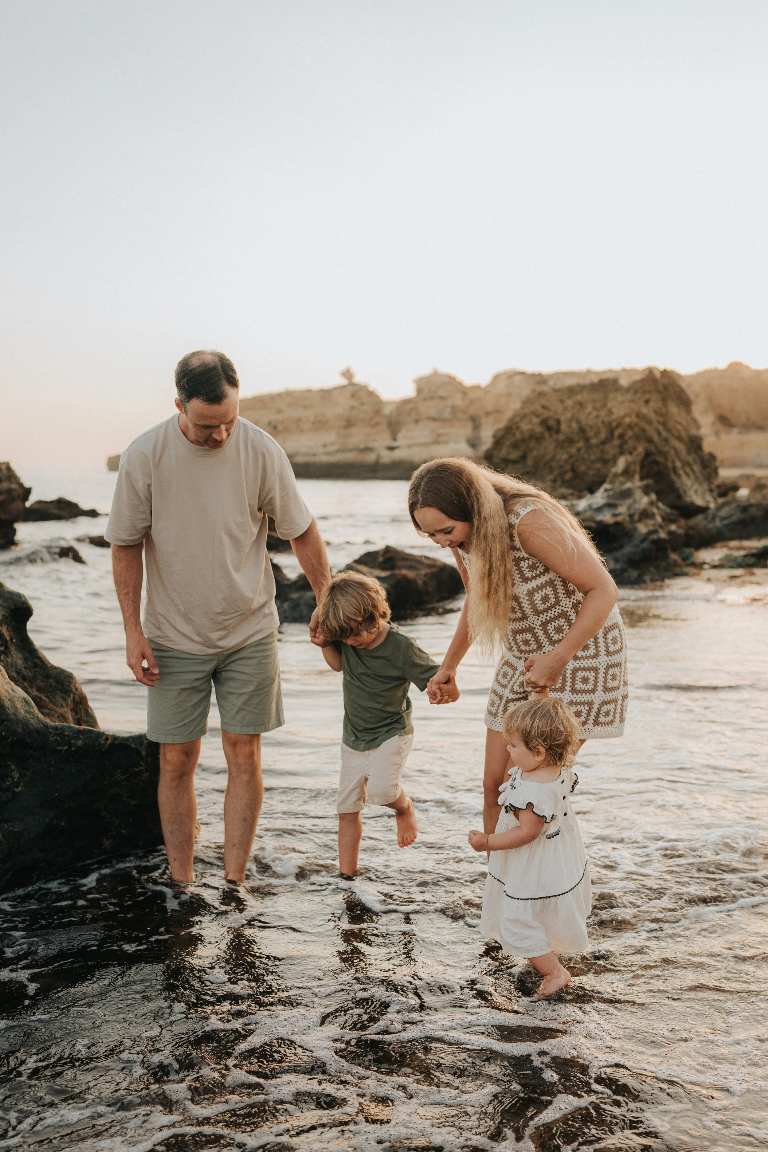 A family of five with two children and two adults enjoying a day at the beach, wading through shallow water with rocky formations and cliffs in the background during sunset.