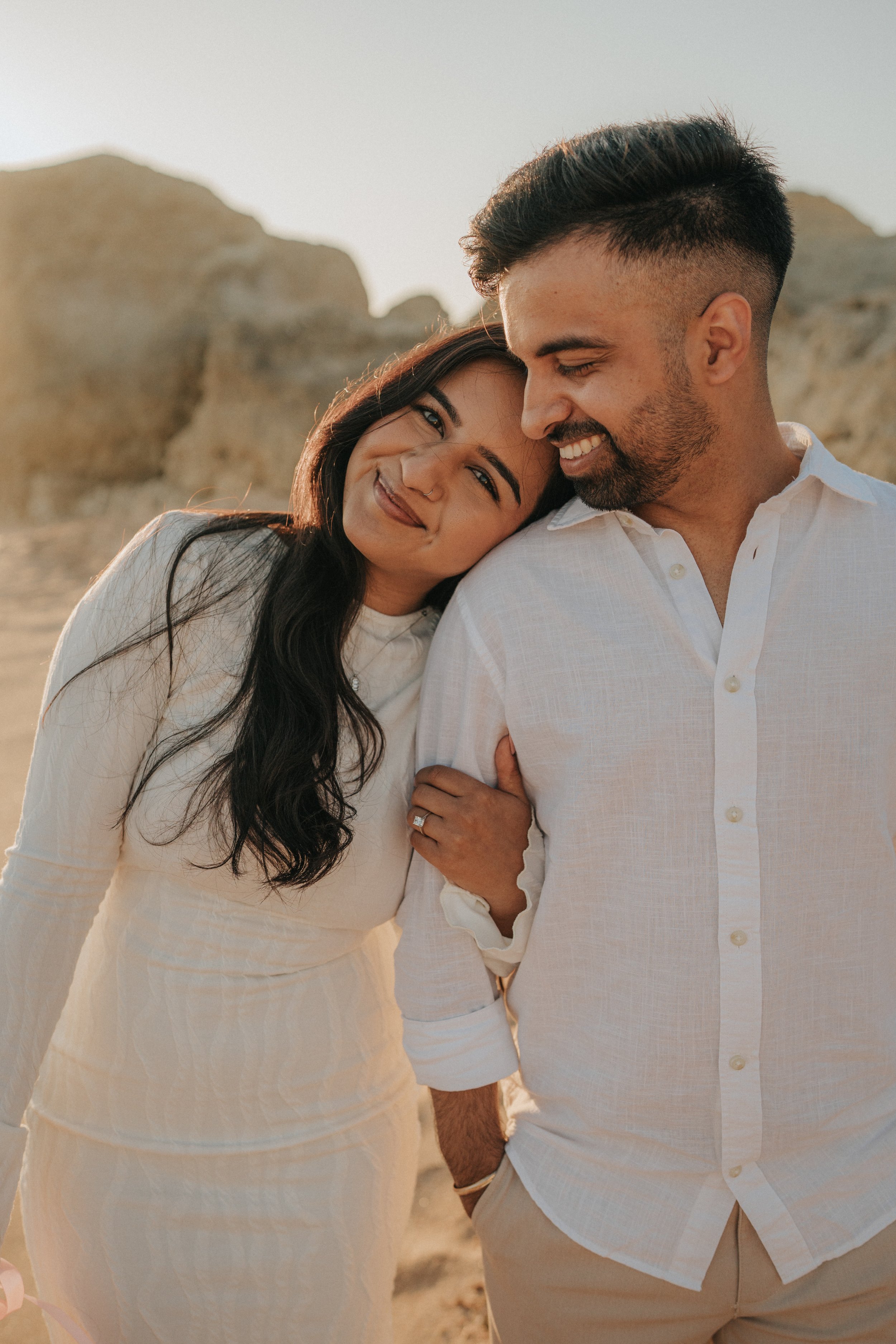 A couple smiling and embracing outdoors with rocky landscape in background, dressed in light-colored clothing.