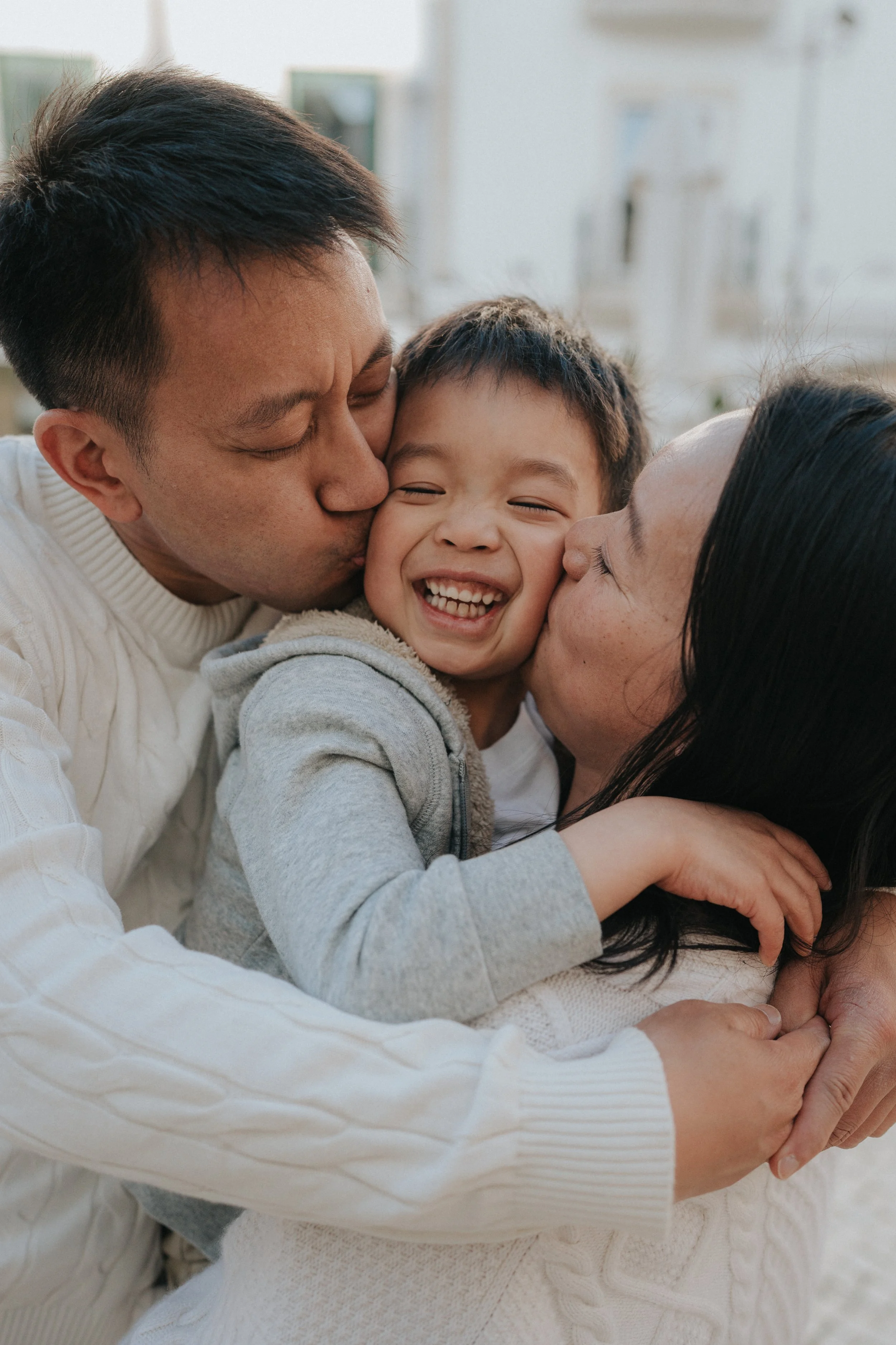 A family of three, including a father, mother, and young boy, sharing a joyful moment with the father kissing the boy and the mother kissing the boy's cheek, outdoors on a bright day.