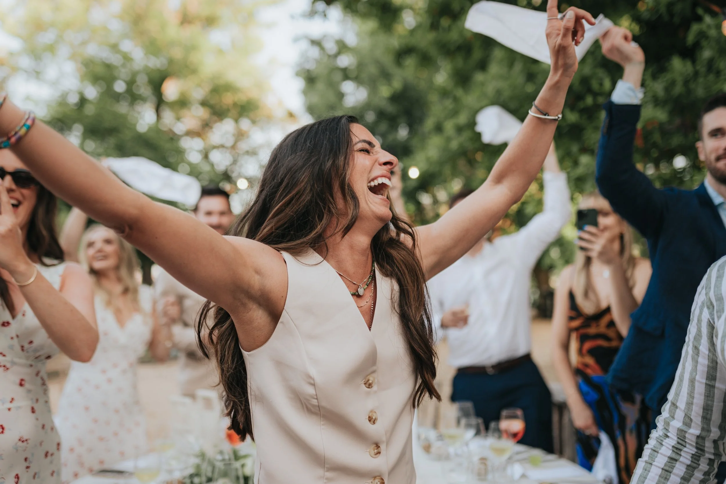 A woman with long brown hair is joyfully raising her arms at an outdoor gathering, surrounded by smiling friends and family, with trees and sunlight in the background.