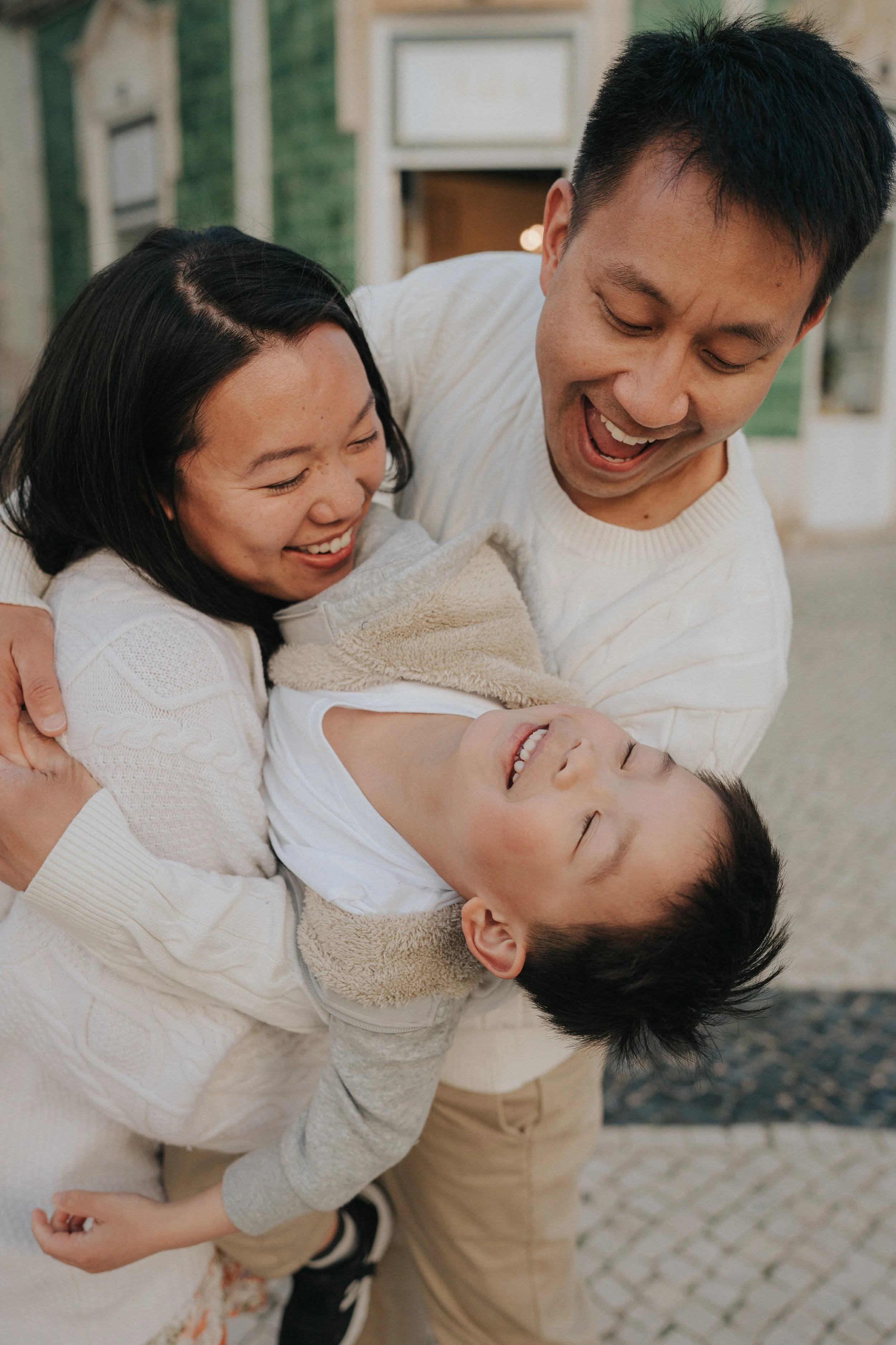 A family of three children and two adults playing and laughing outdoors on a cobblestone path with a house and window in the background.