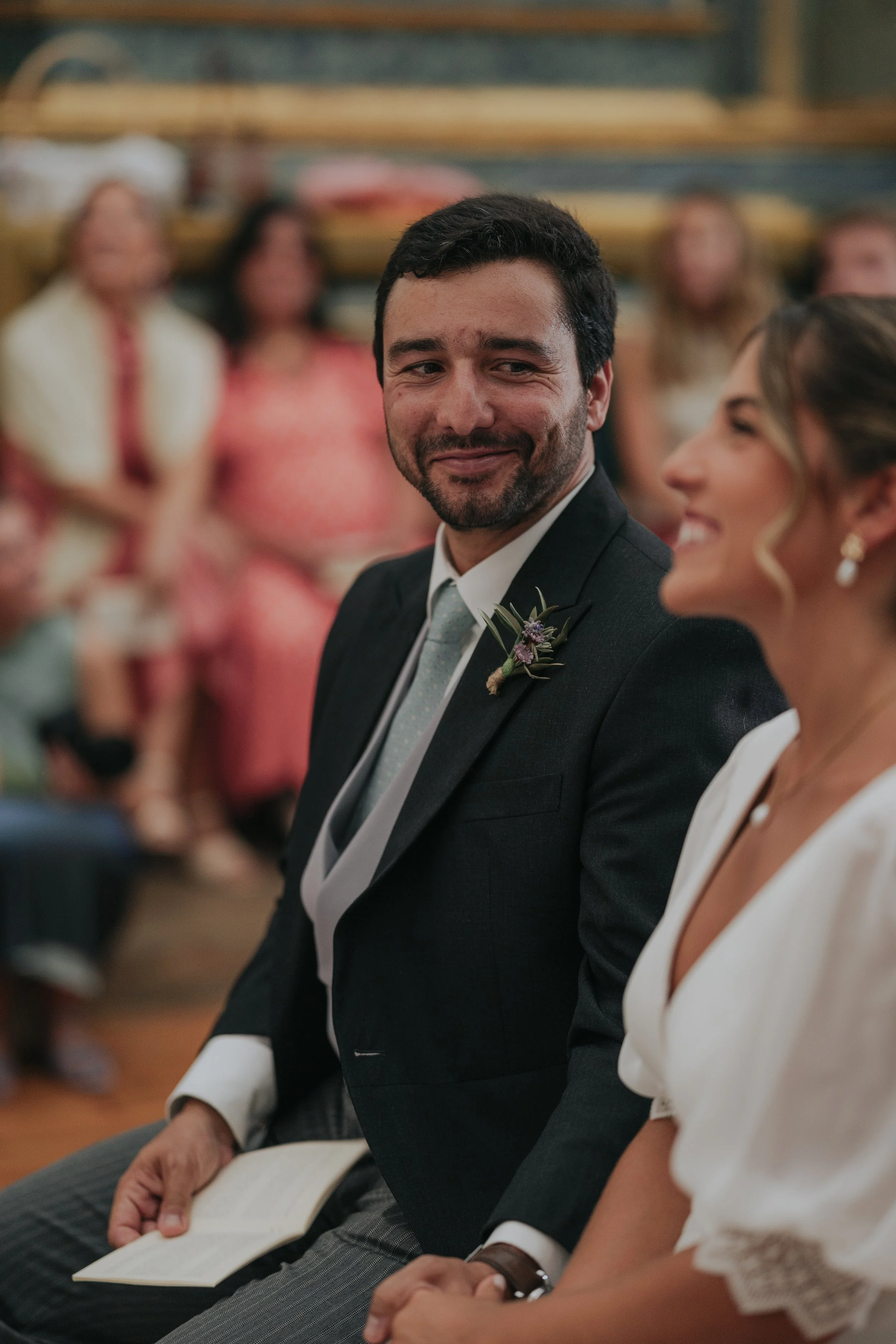 A groom in a dark suit with a boutonniere, sitting next to a woman in a white dress, both smiling, surrounded by guests at a wedding ceremony.