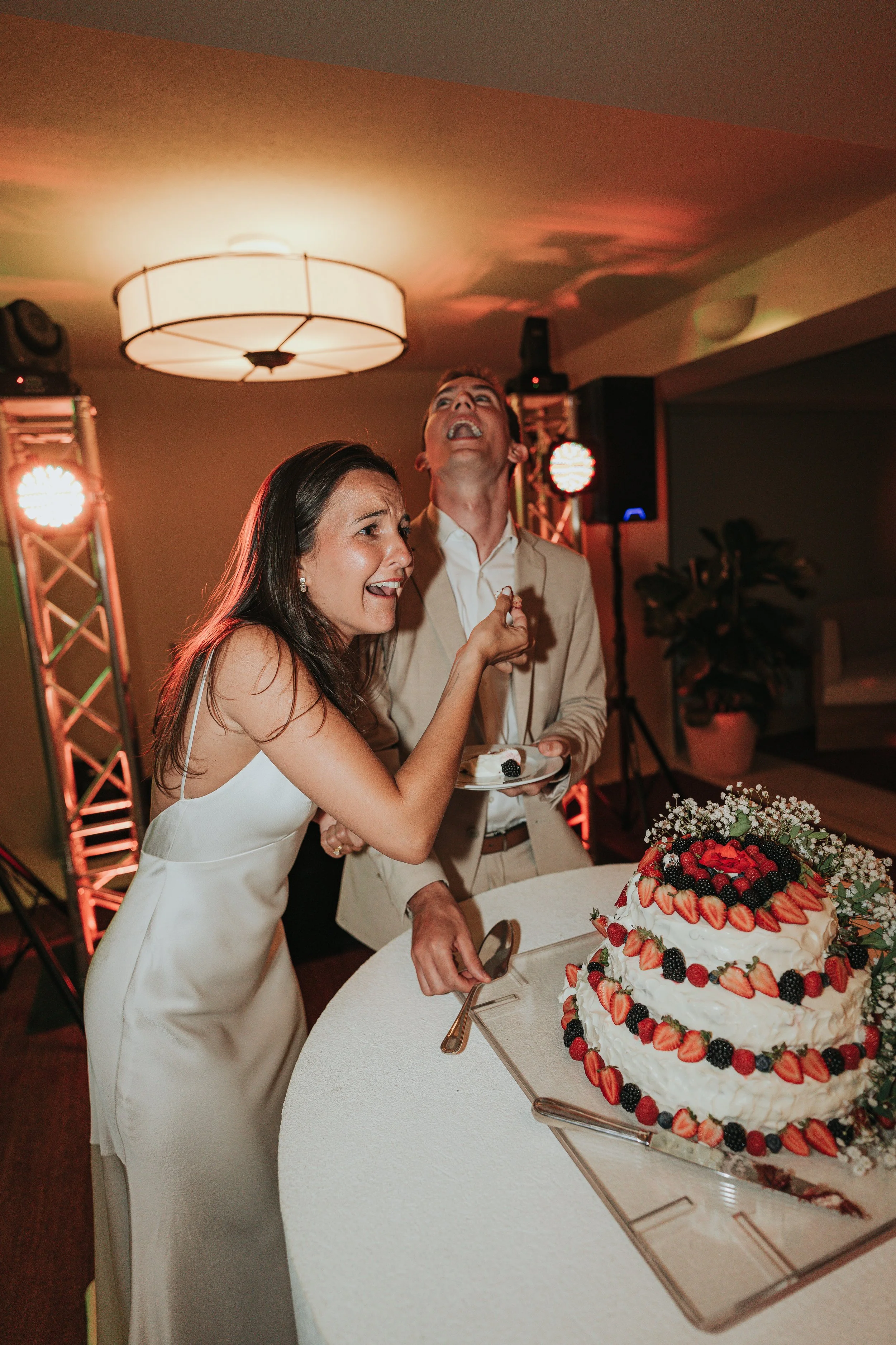 A bride and a groom smiling and laughing as they prepare to cut a wedding cake decorated with strawberries and mixed berries, at a wedding reception.