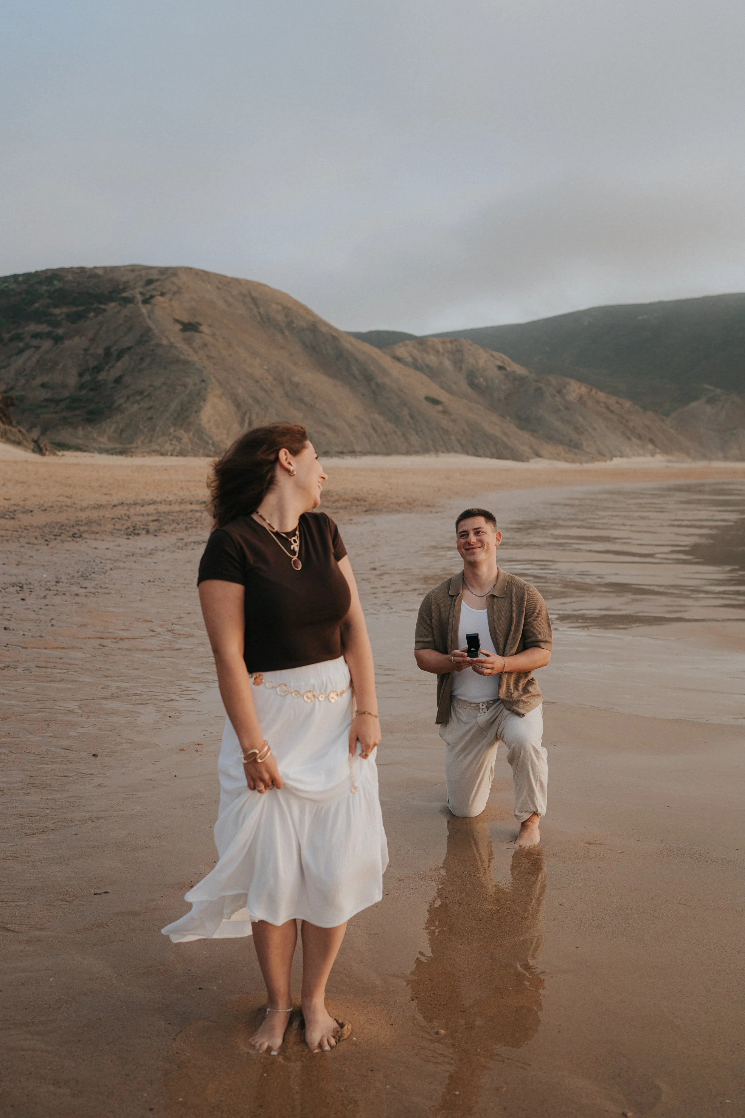 A woman with long brown hair in a black top and white skirt, and a man kneeling in the shallow water holding a phone, at the beach with sandy shore and green hills in the background.