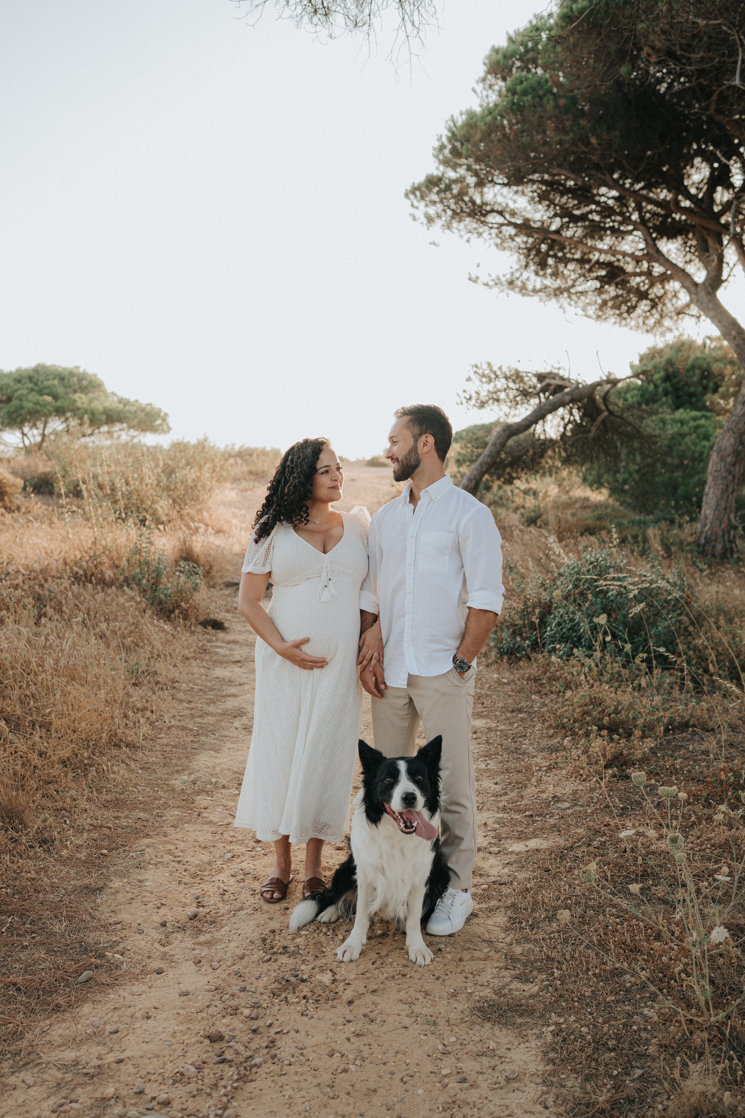 A pregnant woman and a man stand together on a dirt trail in a natural, dry landscape with trees. The woman wears a white dress, and the man wears a white shirt and beige pants. A black and white dog is sitting in front of them with its tongue out.