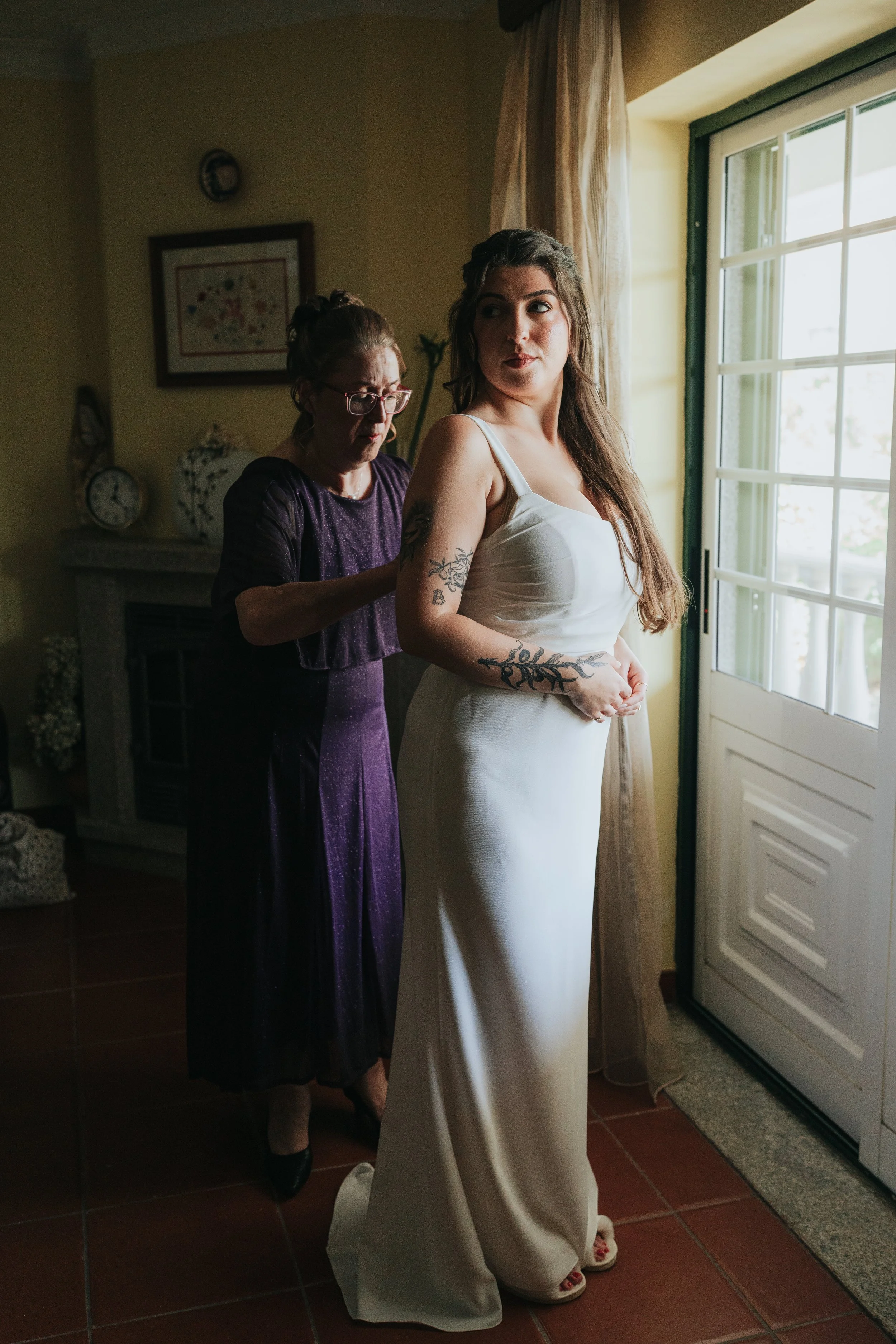 A bride in a white dress with tattoos on her arms having her dress adjusted by an older woman in a purple dress. They are by a sliding glass door with beige curtains in a warmly lit room.