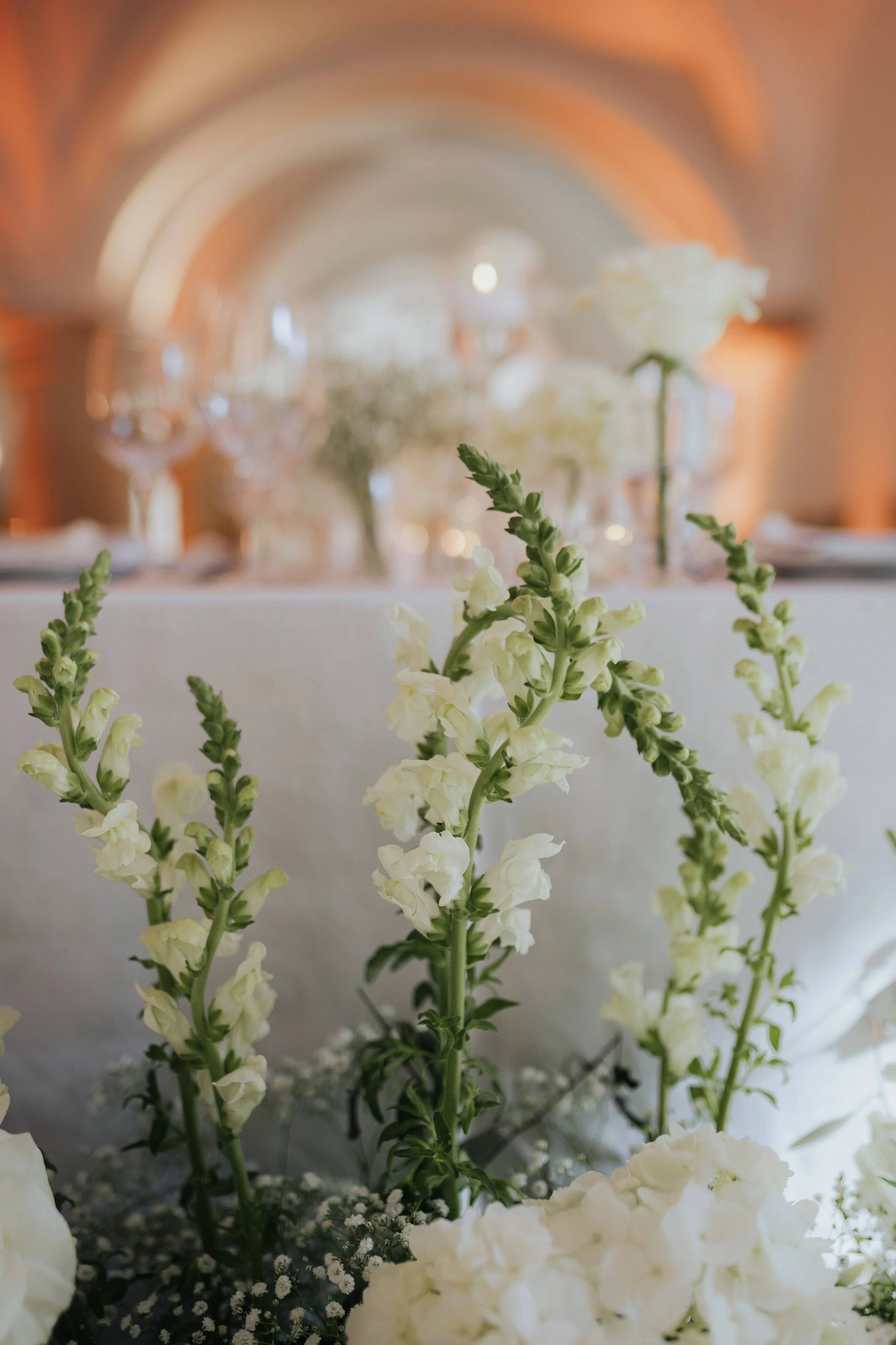 White and green flower arrangement, with white flowers in the foreground and a blurred background featuring a chandelier and warm lighting.
