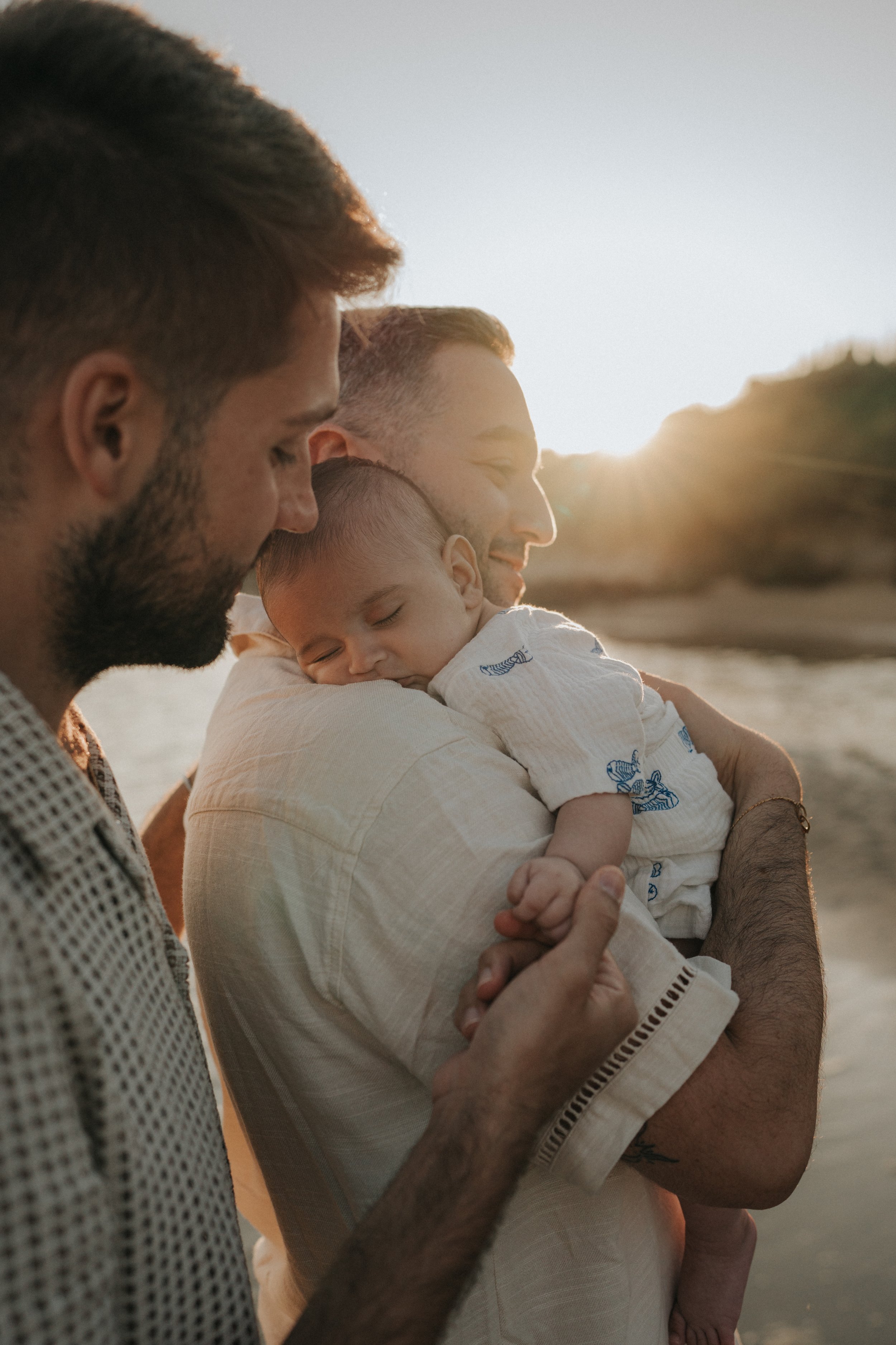 Three men with a baby at sunset outside, close-up shot showing their side profiles, warm sunlight background.
