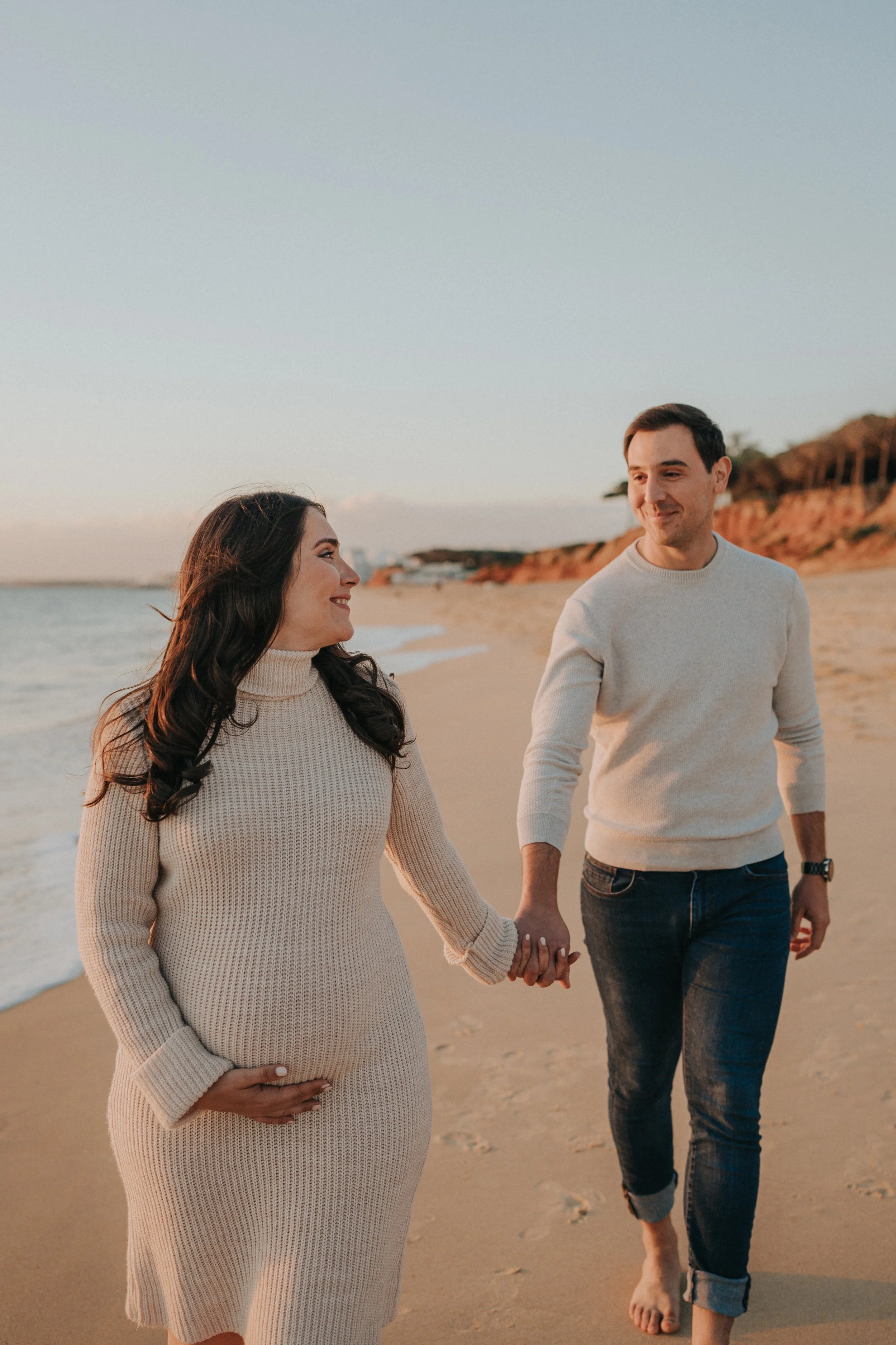 A pregnant woman and a man walk on the beach holding hands during sunset, smiling and looking at each other.