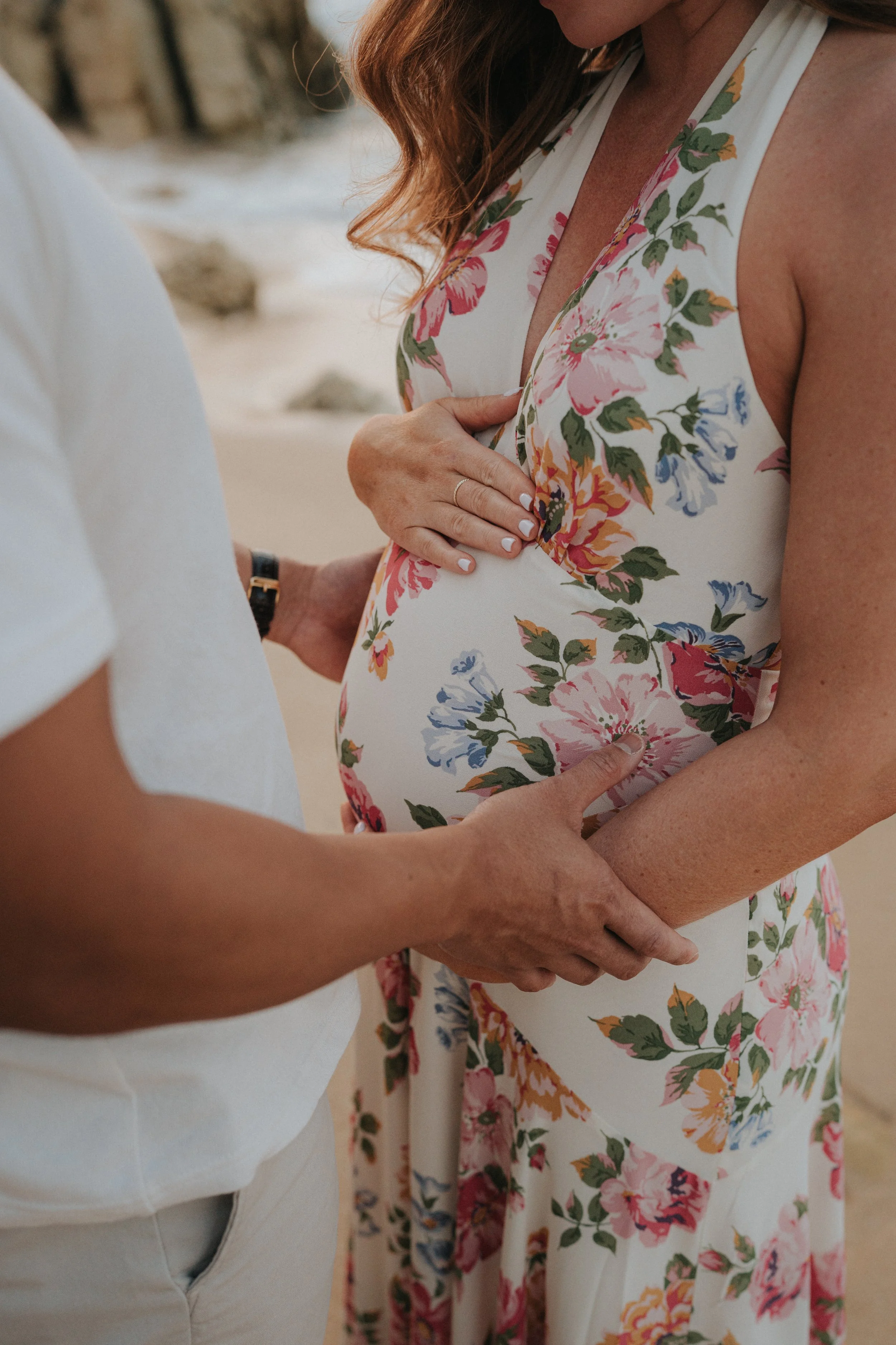 A woman wearing a floral dress and a person with clasped hands holding her baby bump.