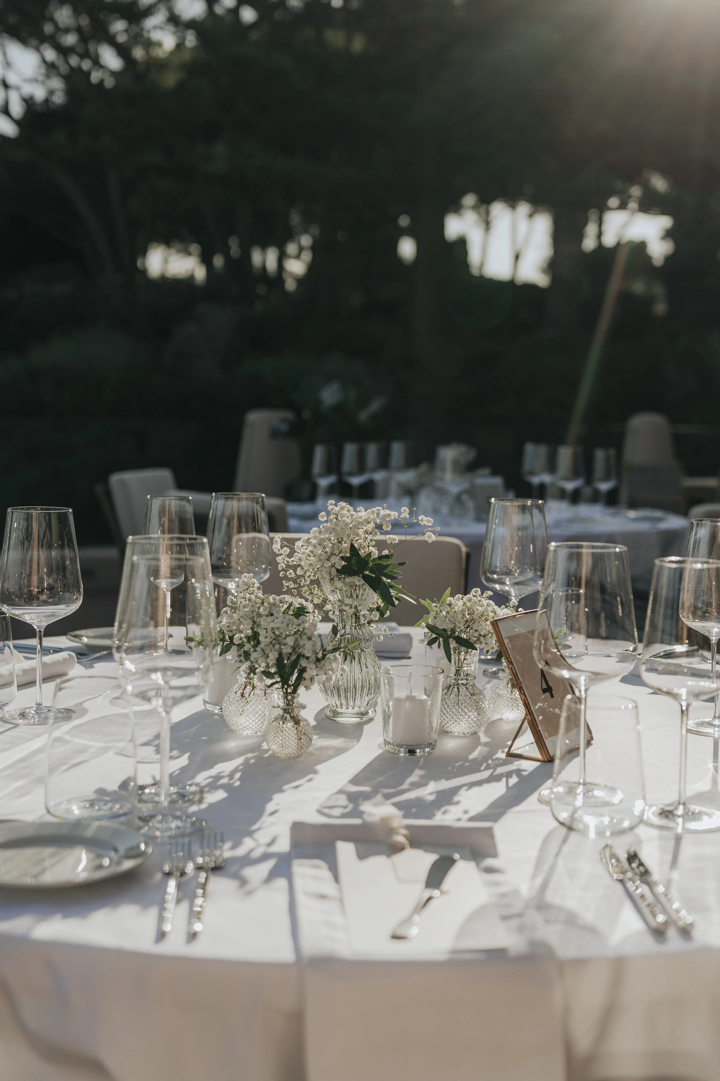 A table set for a formal event with white tablecloth, multiple wine glasses, silverware, and floral centerpieces with white flowers in glass vases. There is a small table number card in a copper frame.