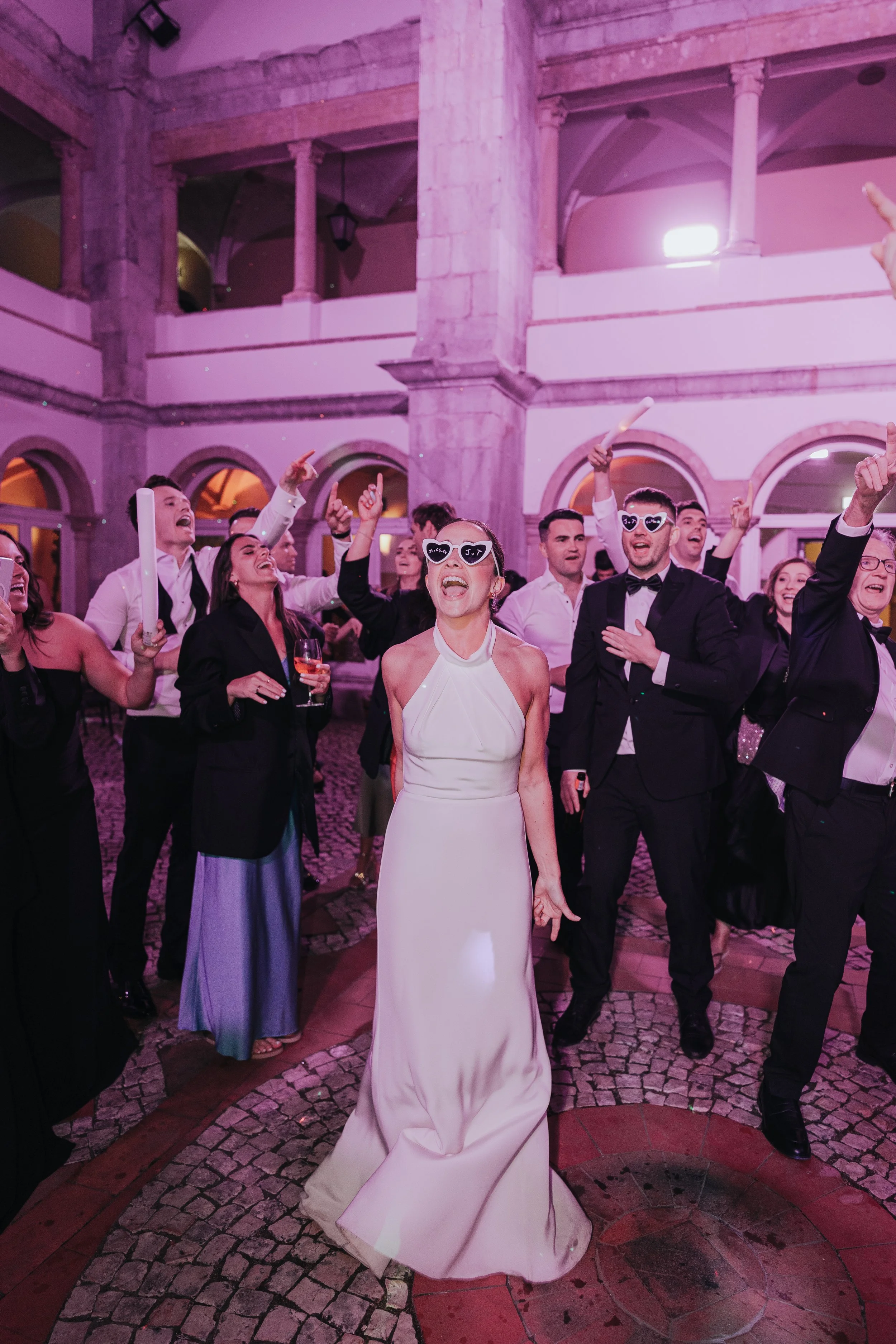 A woman in a white halter dress with sunglasses is dancing and smiling at a lively party, surrounded by people in formal attire inside a grand, historic building with pink and purple lighting.