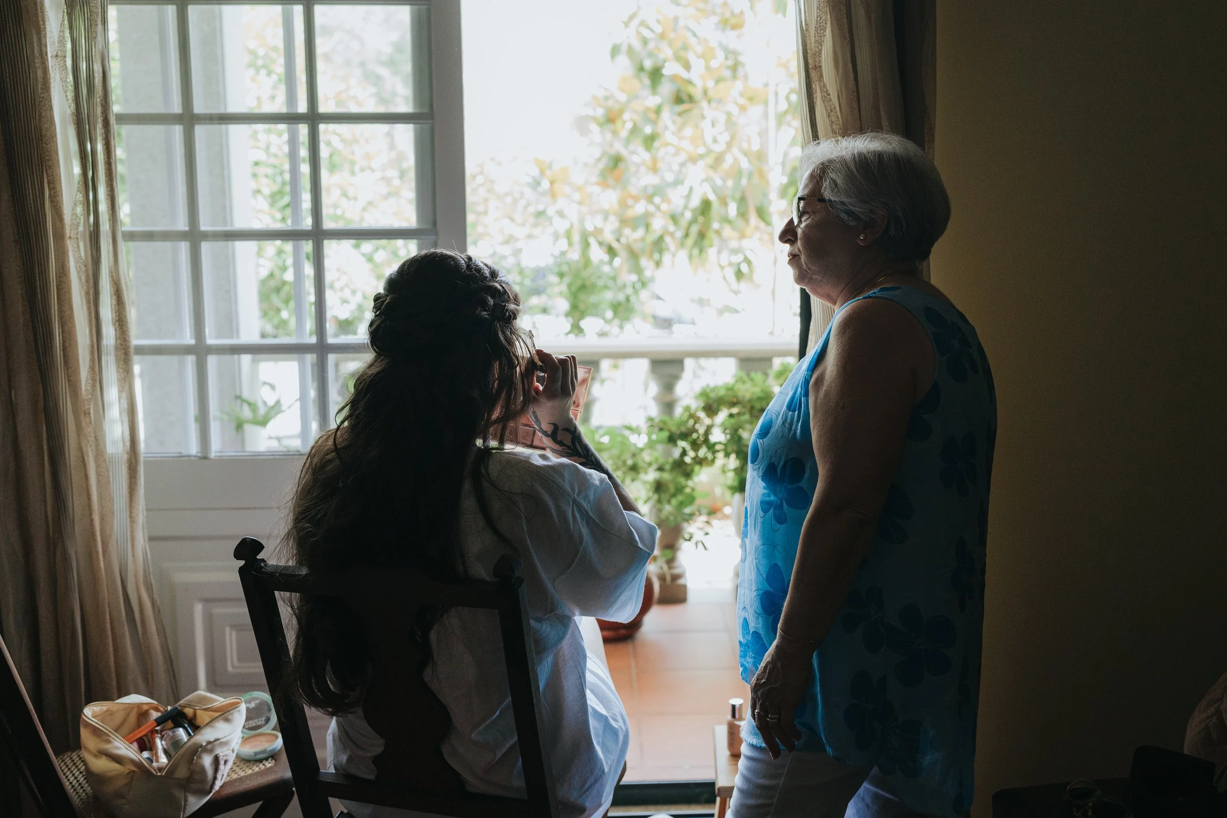 An elderly woman with gray hair and glasses standing and talking to a younger woman with long hair in a wheelchair, near a window with sunlight coming through, in a cozy indoor setting.