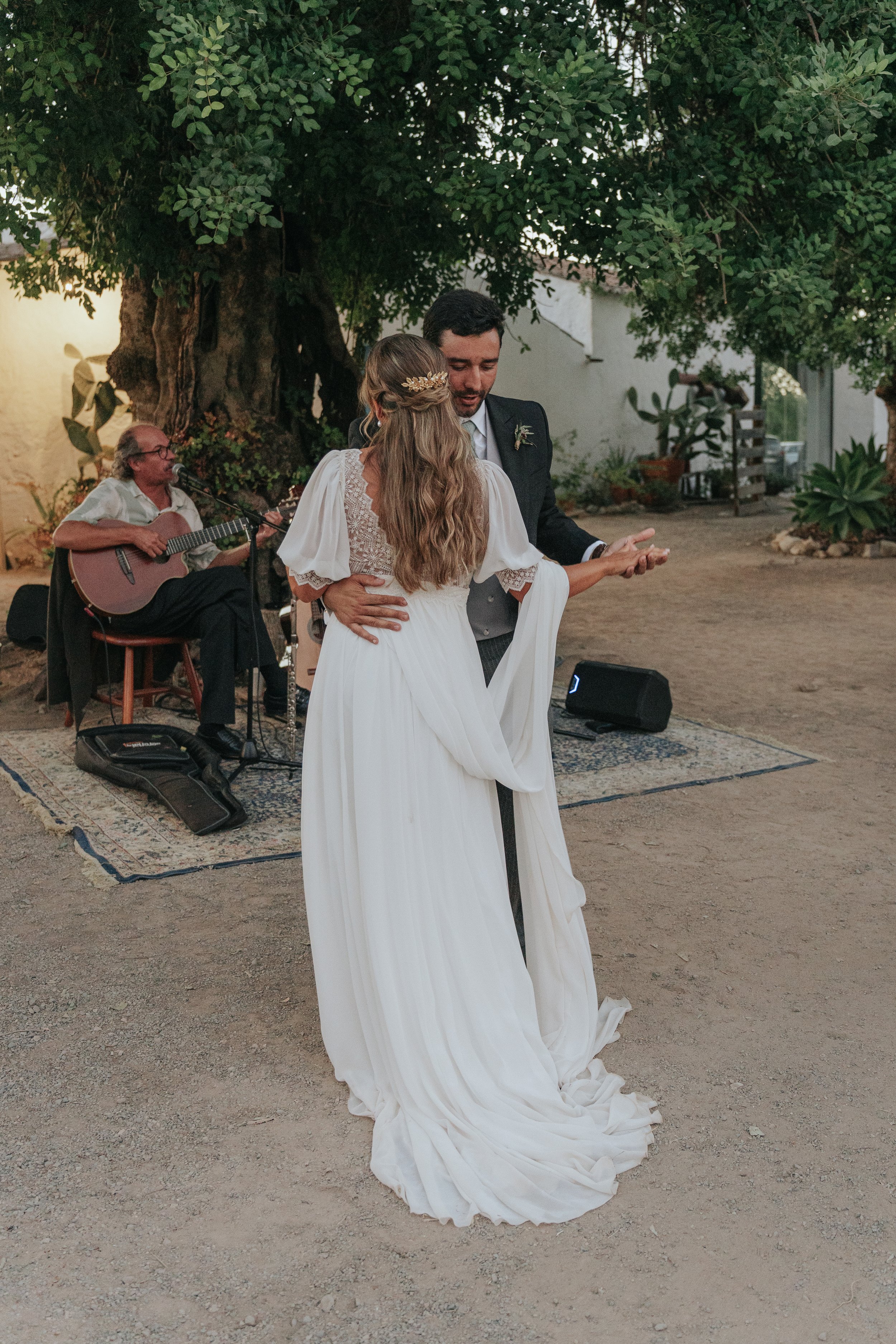 A bride and groom sharing a dance at an outdoor wedding reception beneath a large tree, with a musician playing guitar in the background.