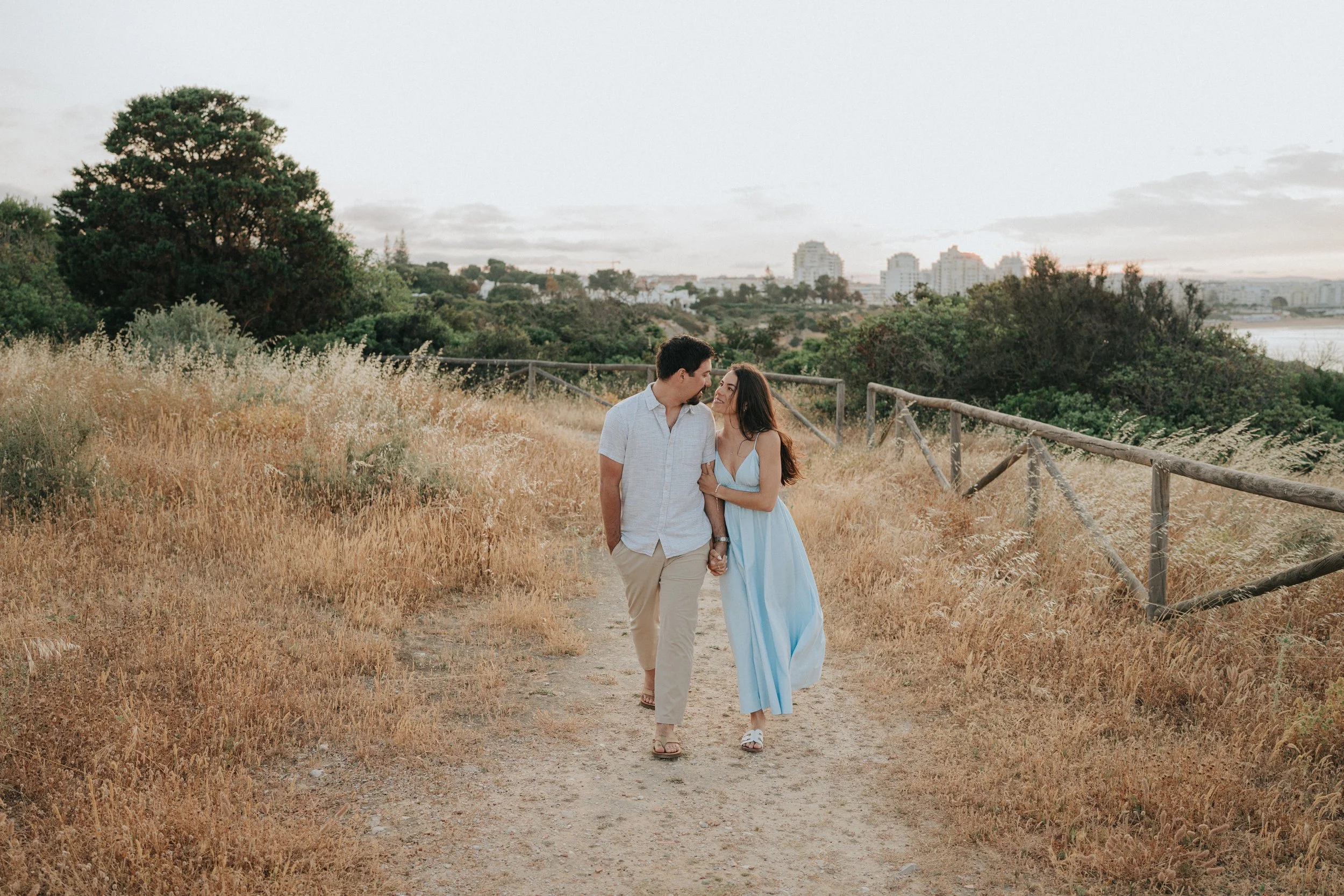A couple walking hand in hand on a dirt trail in a grassy field with trees and city buildings in the distance during sunset.
