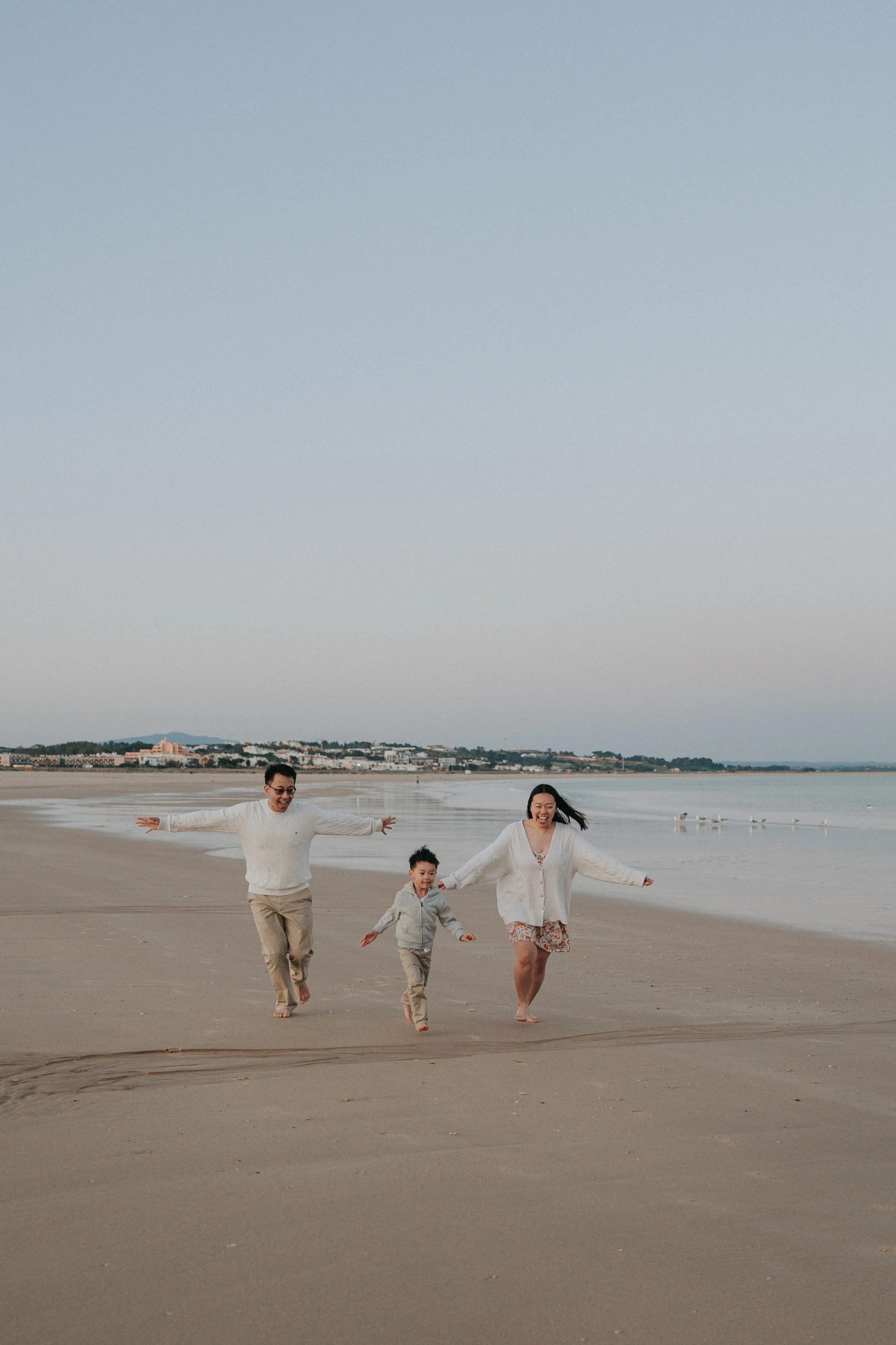 A family of four, including a man, woman, and two children, running happily on a beach with their arms and hands outstretched, with the ocean and a distant city skyline in the background.