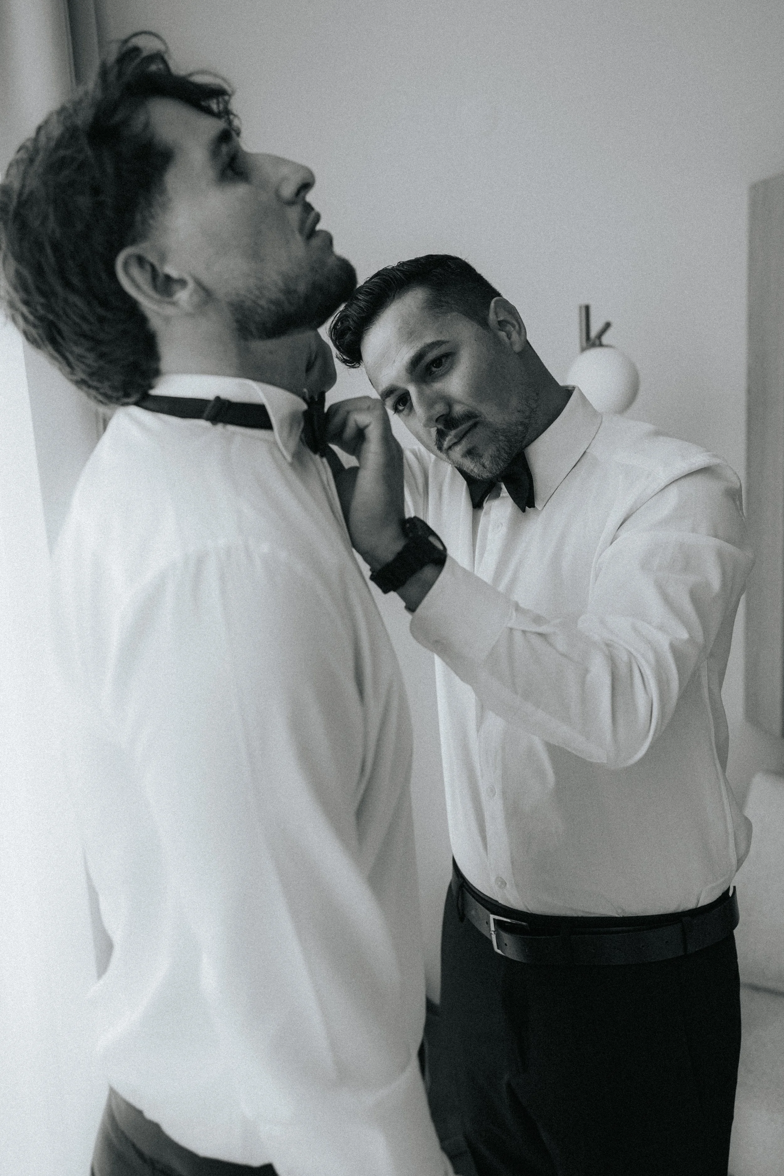 Two men dressing formally in a bedroom, with one adjusting the other's bowtie.