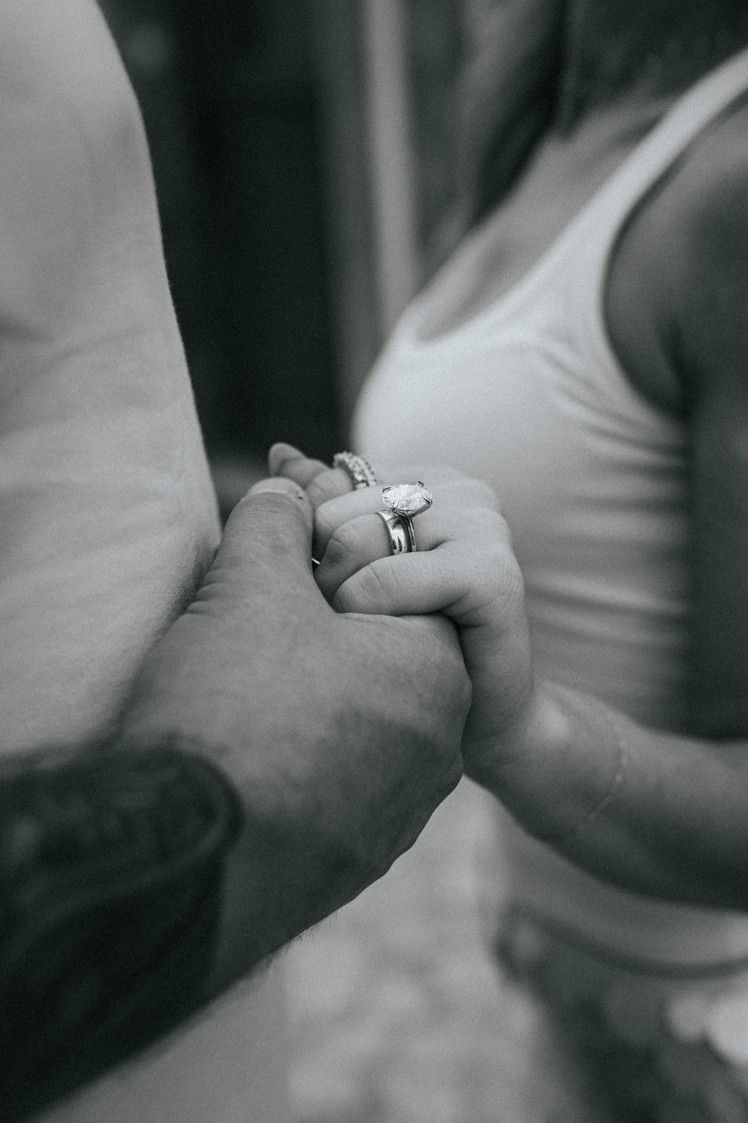Close-up black and white photo of a person holding another person's hand, with one wearing a ring with a large gemstone.