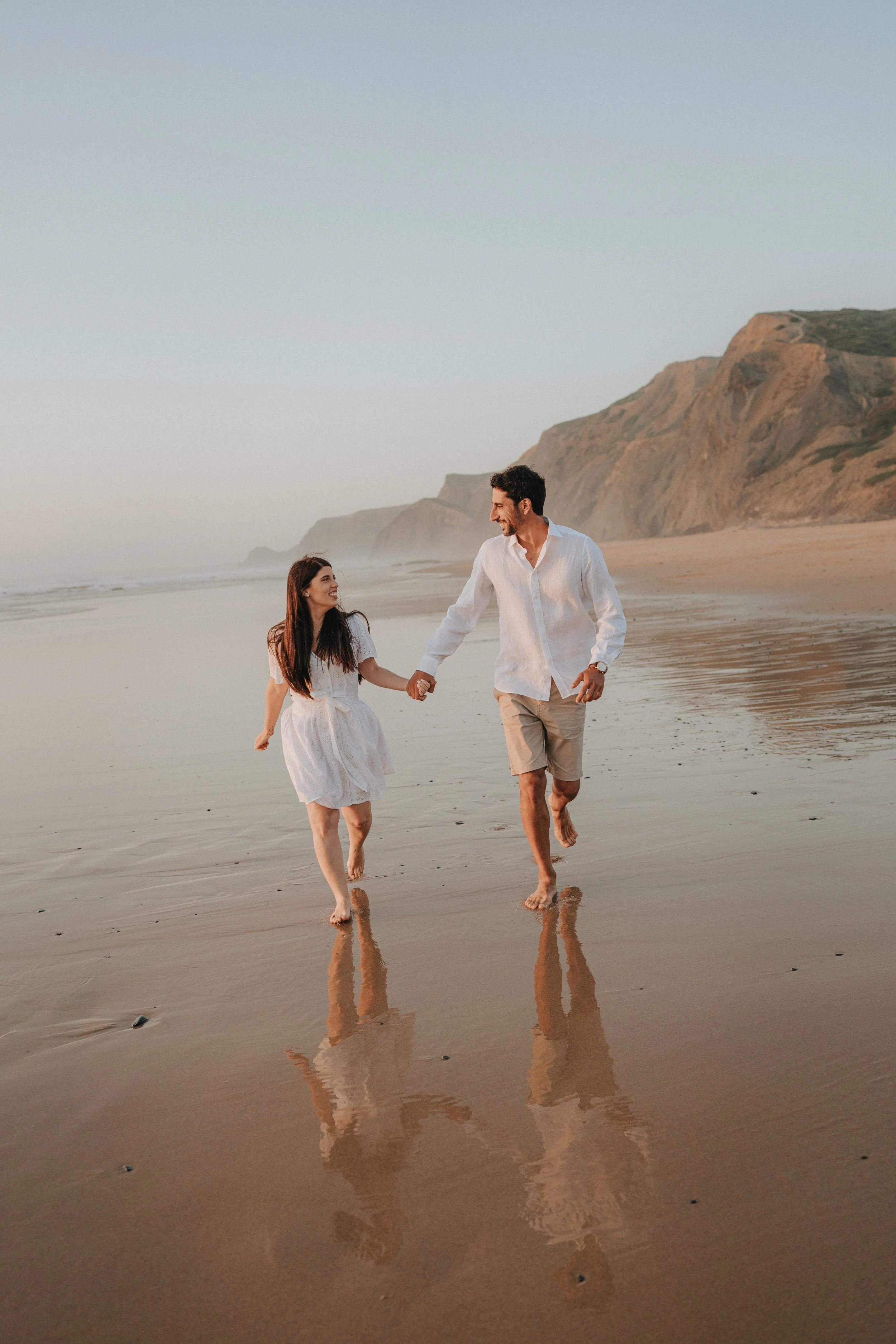 A couple holding hands walking barefoot on a beach at sunset, with cliffs in the background and their reflections visible on the wet sand.