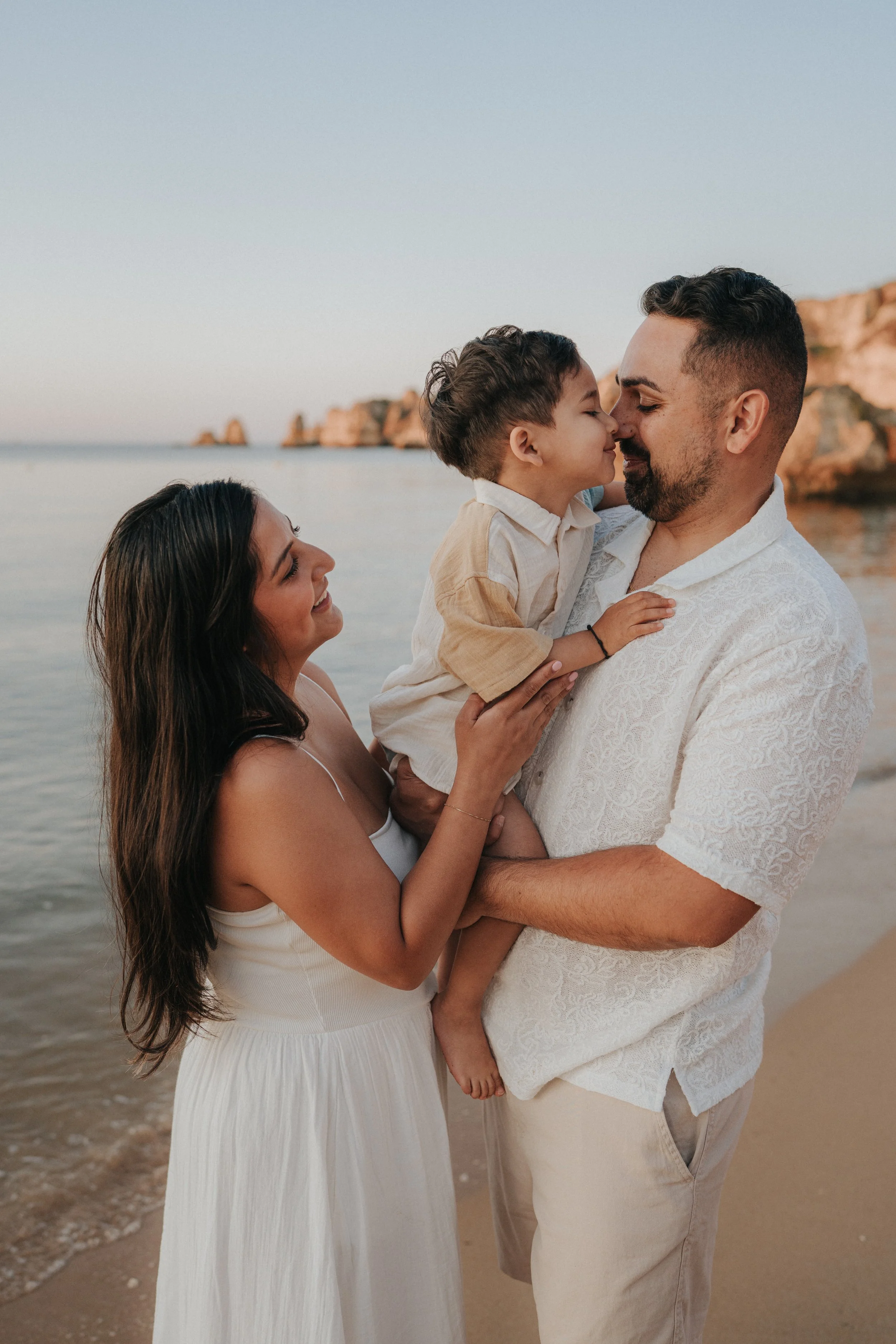 A family of three on a beach at sunset, with the father holding a young boy and the mother smiling at them. The family is close and smiling, with rocks and the ocean in the background.