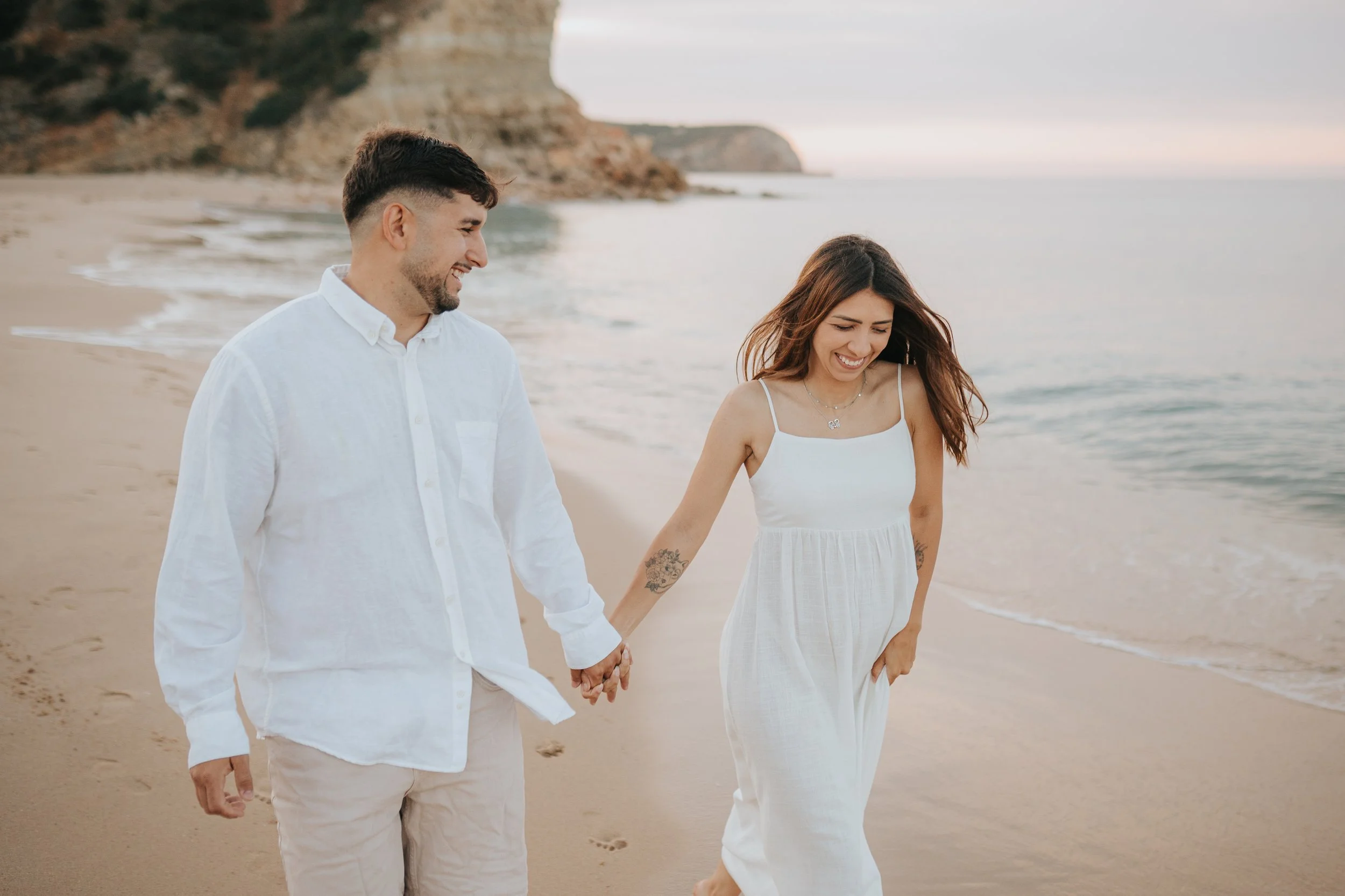 A smiling man and woman holding hands and walking on the beach, dressed in white clothes.
