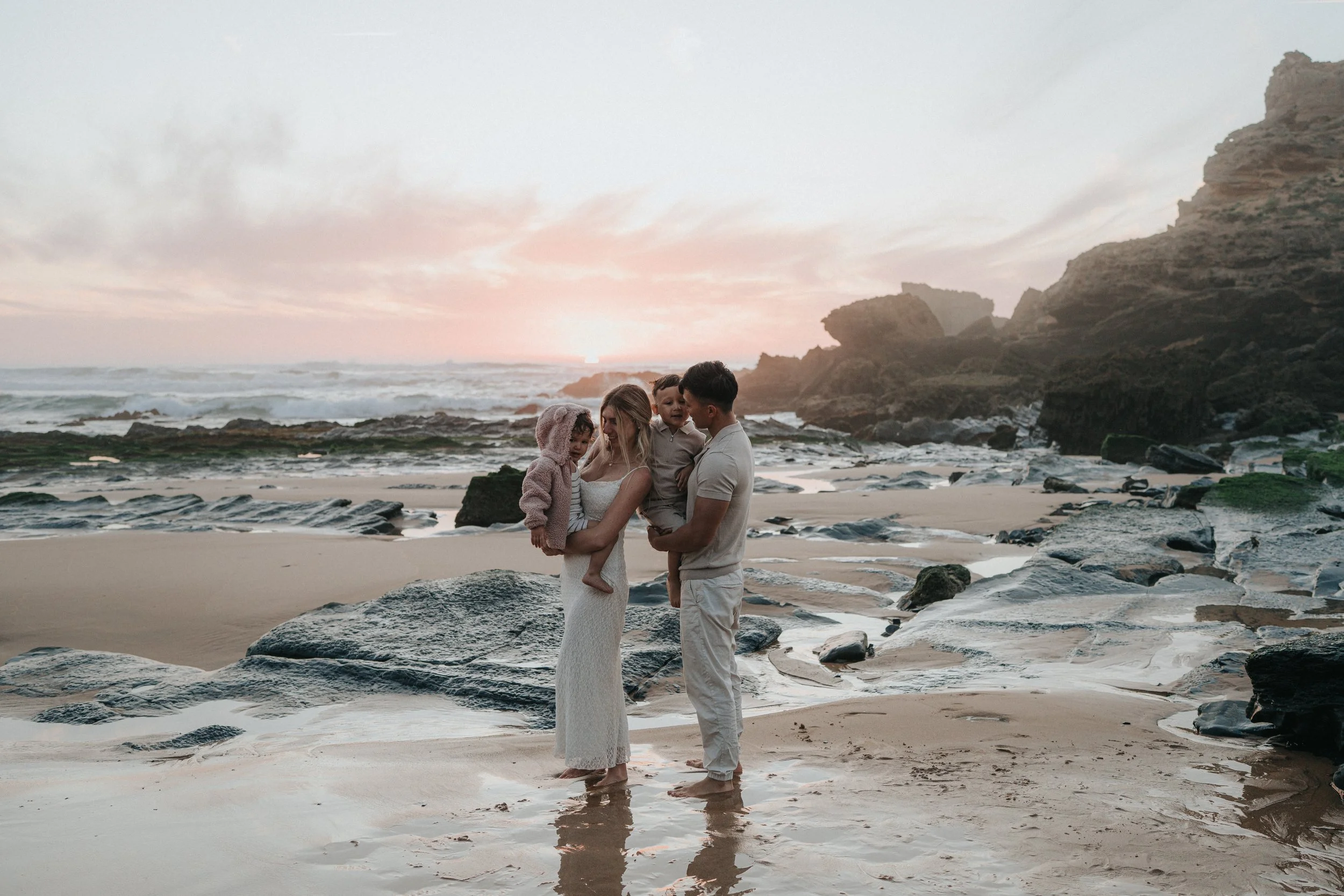 A family of four standing barefoot on a rocky beach at sunset, near the ocean, with cliffs in the background.