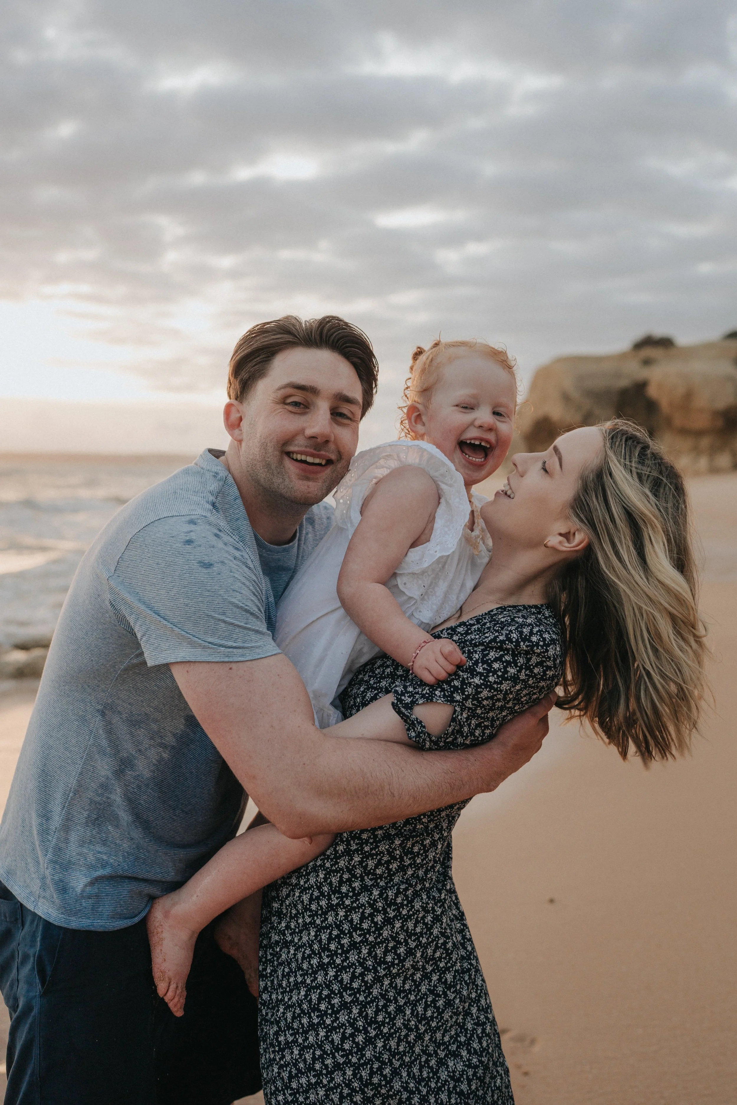 A family of three at the beach during sunset. The man is smiling and holding a young girl, who is laughing. The woman is looking at the girl with a smile. The background shows the ocean, rocks, and a cloudy sky.