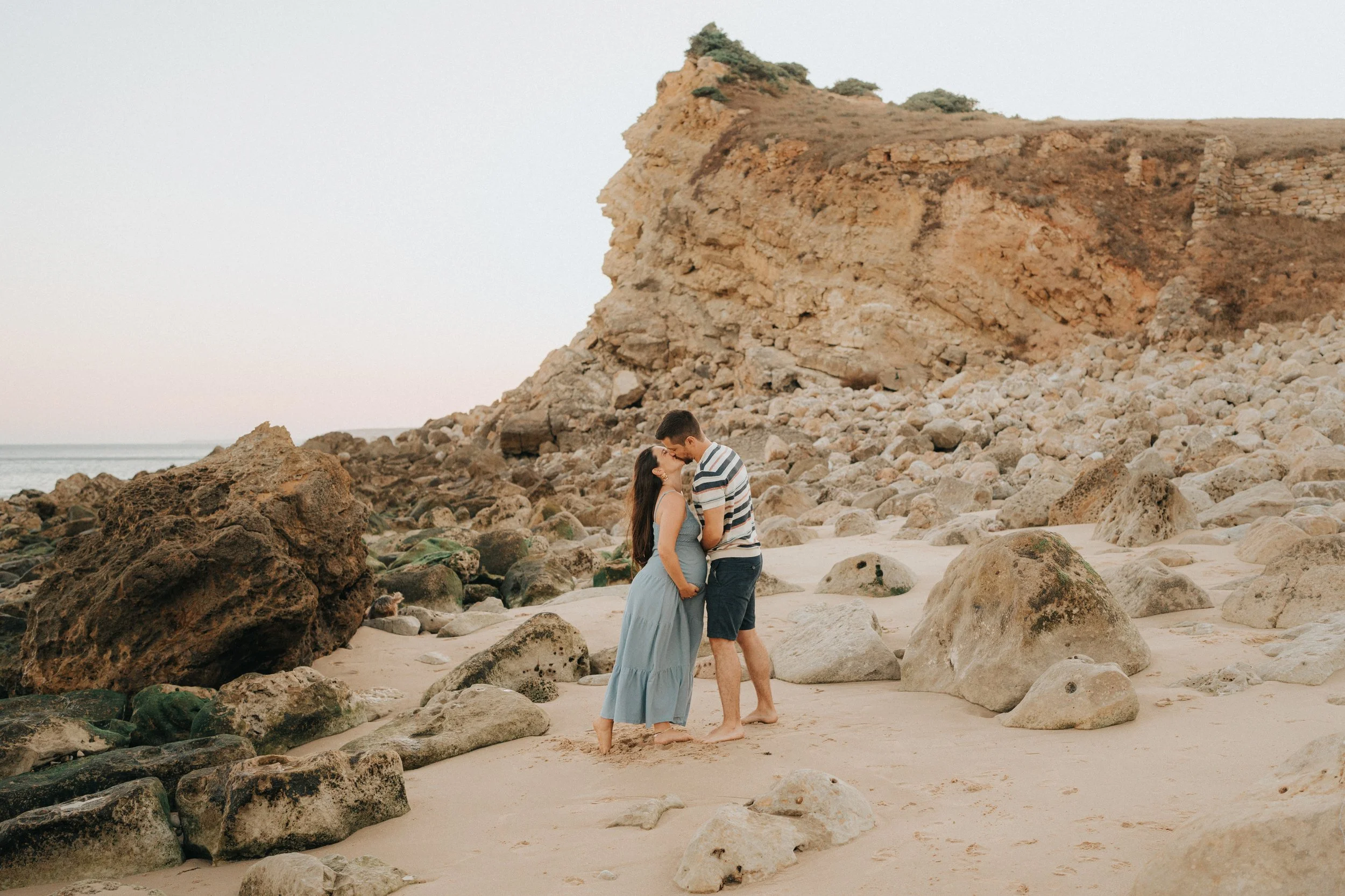 A couple stands on a rocky beach, holding hands and leaning in for a kiss, with large rocks and a cliff in the background.