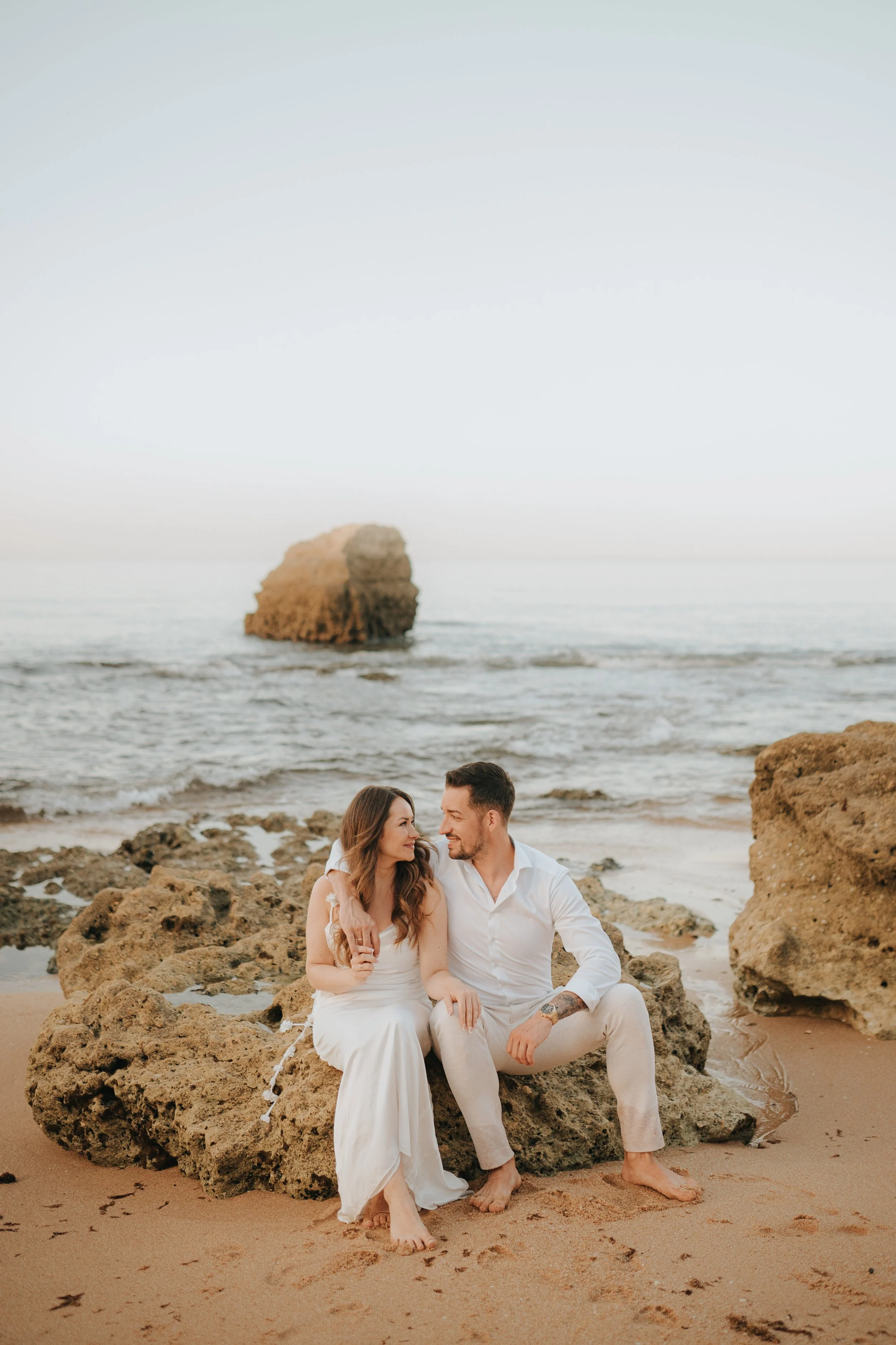 A couple sitting on rocks at the beach, gazing into each other's eyes with the ocean and a large rock formation in the background.