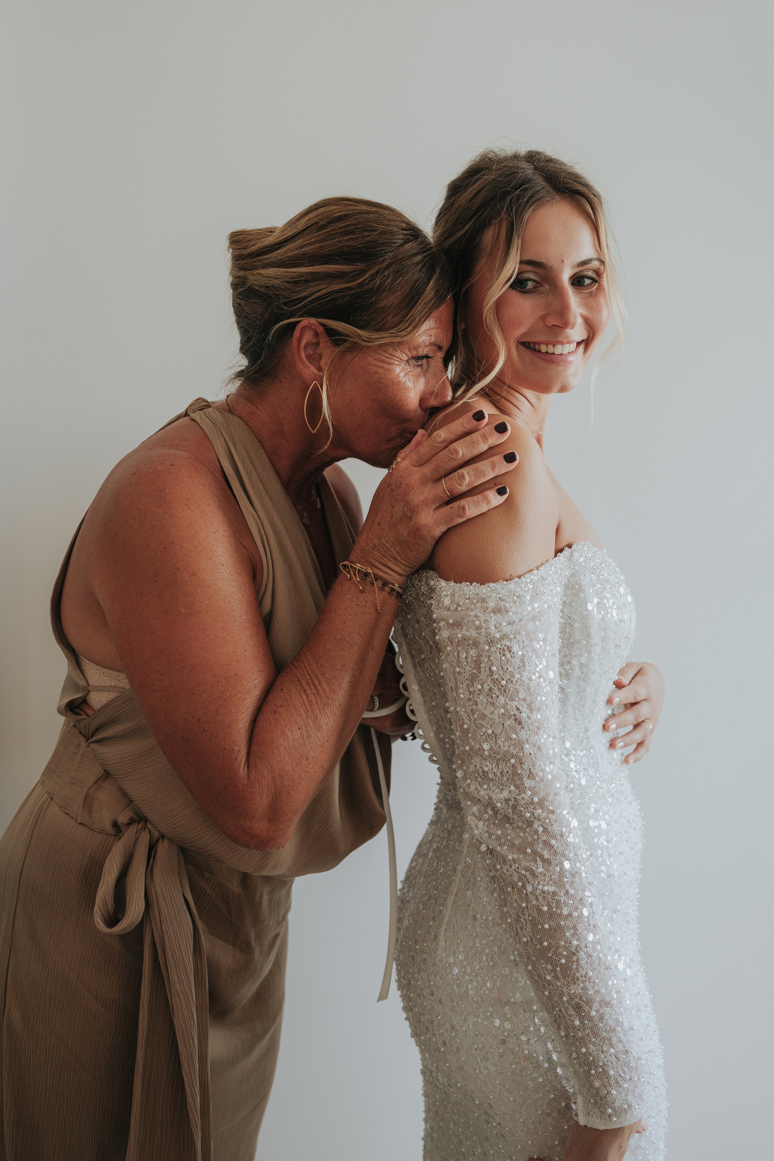An older woman with brown hair, wearing a tan dress, is kissing a young woman on the cheek. The young woman has blonde hair, styled in loose waves, and is wearing a white, off-shoulder, beaded dress. Both women are smiling, and the background is plai