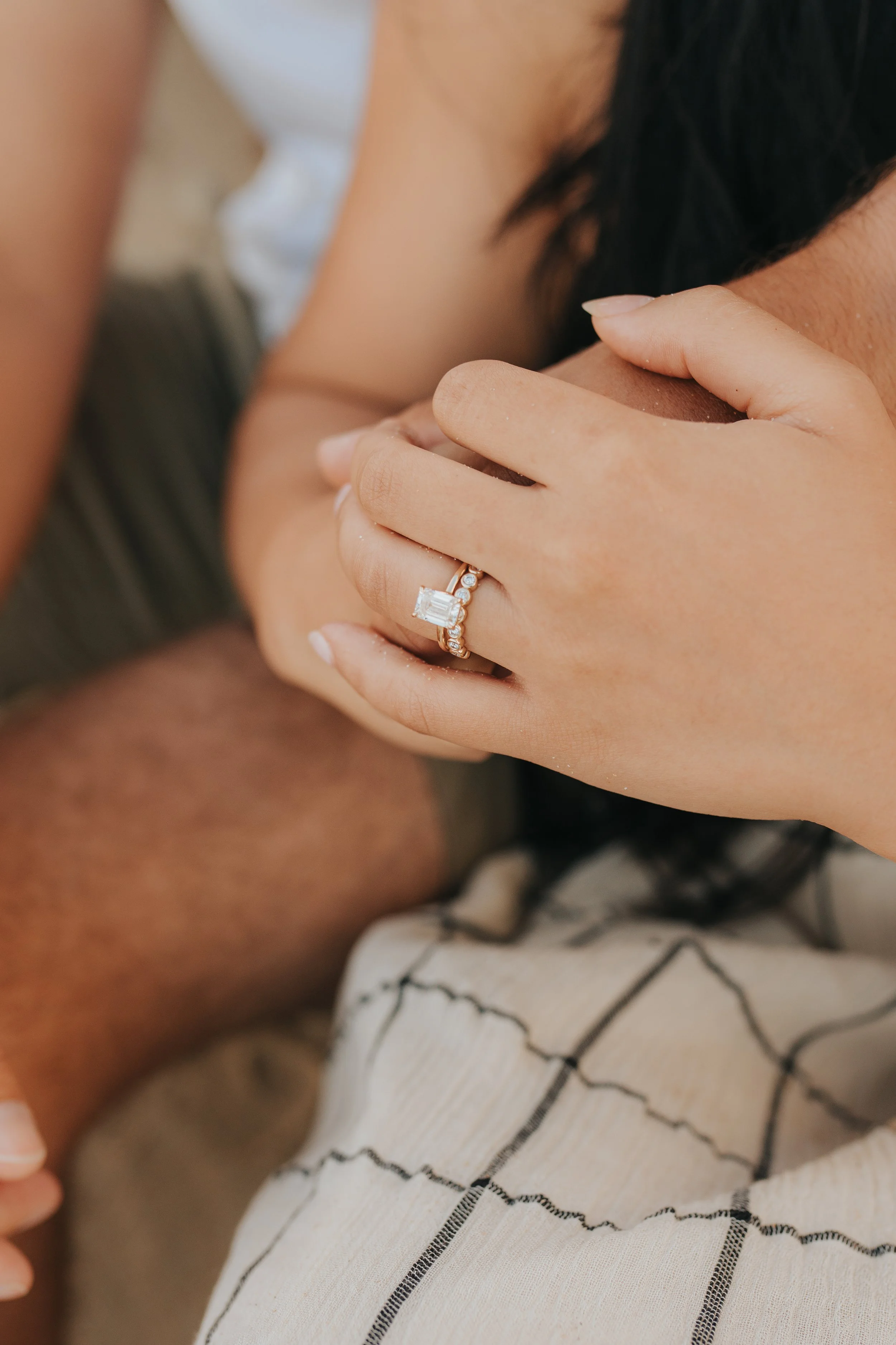Close-up of a woman holding a man's shoulder, wearing a diamond engagement ring and a wedding band, with a patterned fabric in the foreground.