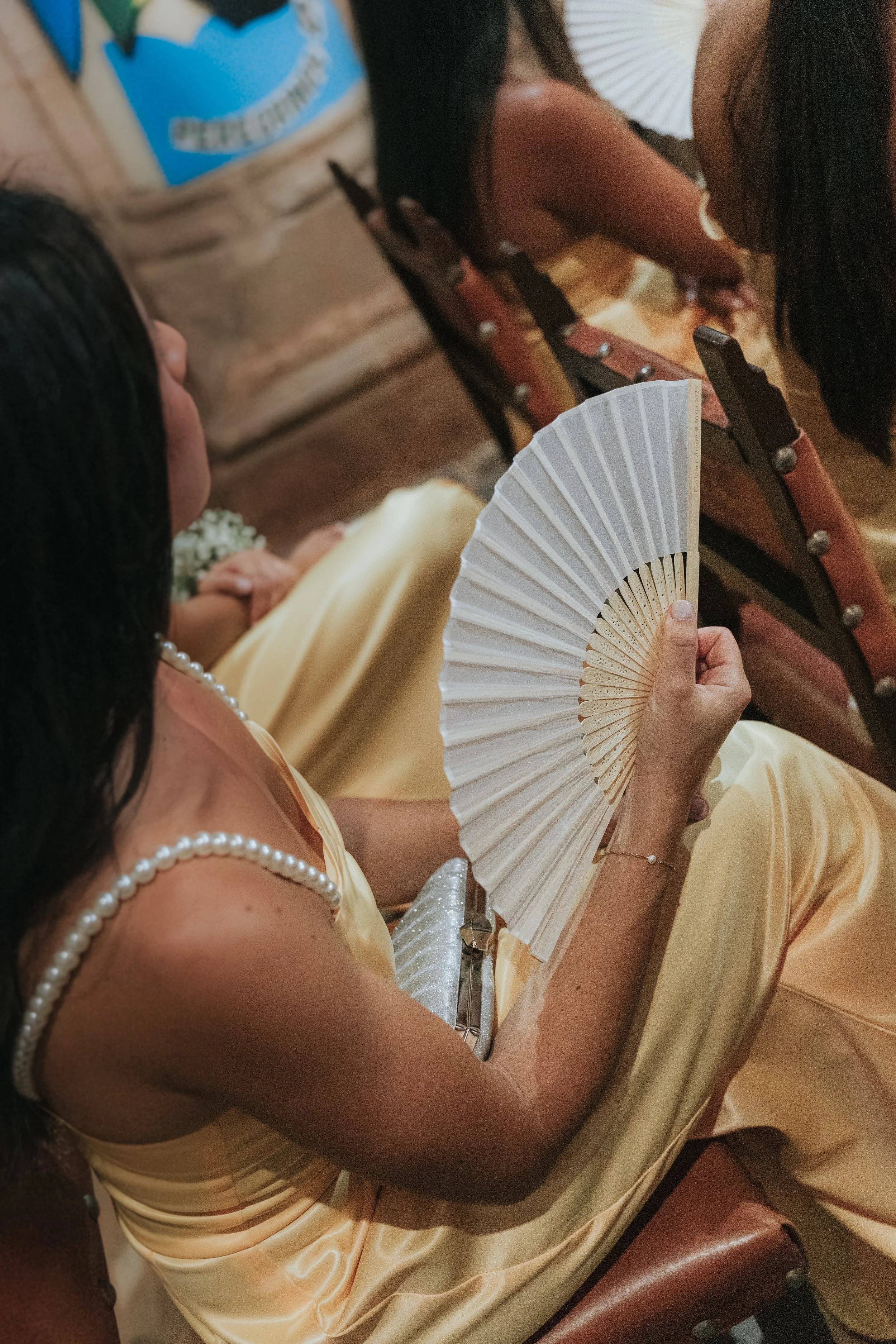 A woman sitting in a row of chairs wearing a yellow dress with pearl details, holding a white folding fan in her hand.