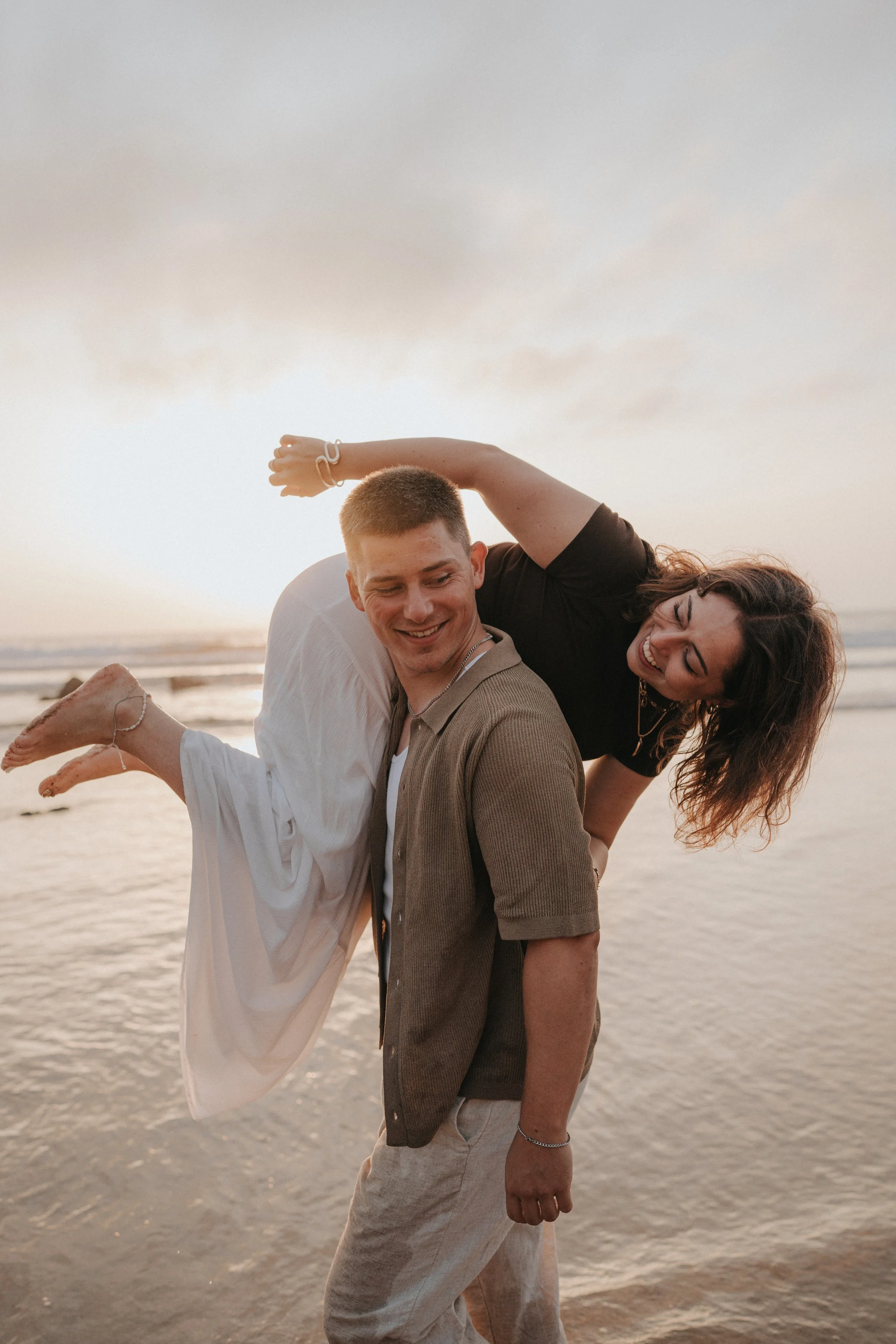 A young man and woman smiling and leaning on each other on the beach during sunset, with the ocean in the background.