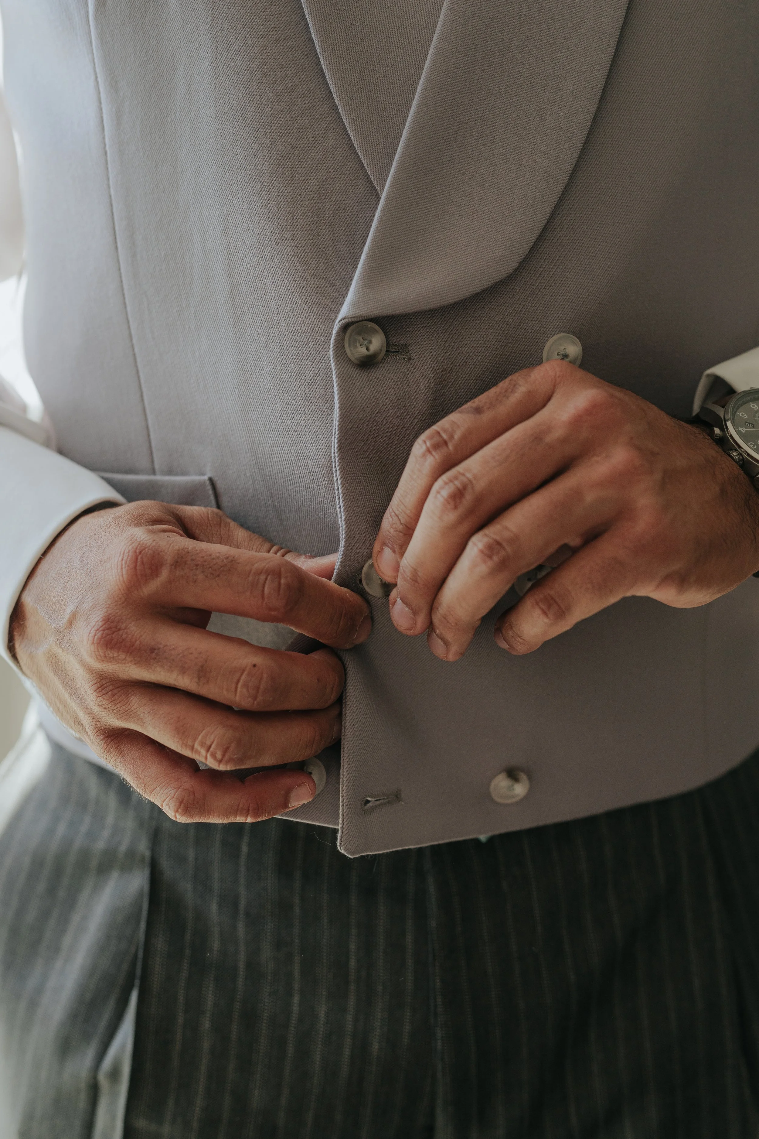 A close-up of a person fastening buttons on a light gray suit jacket, with hands adjusting the buttons and a wristwatch visible on the right wrist.
