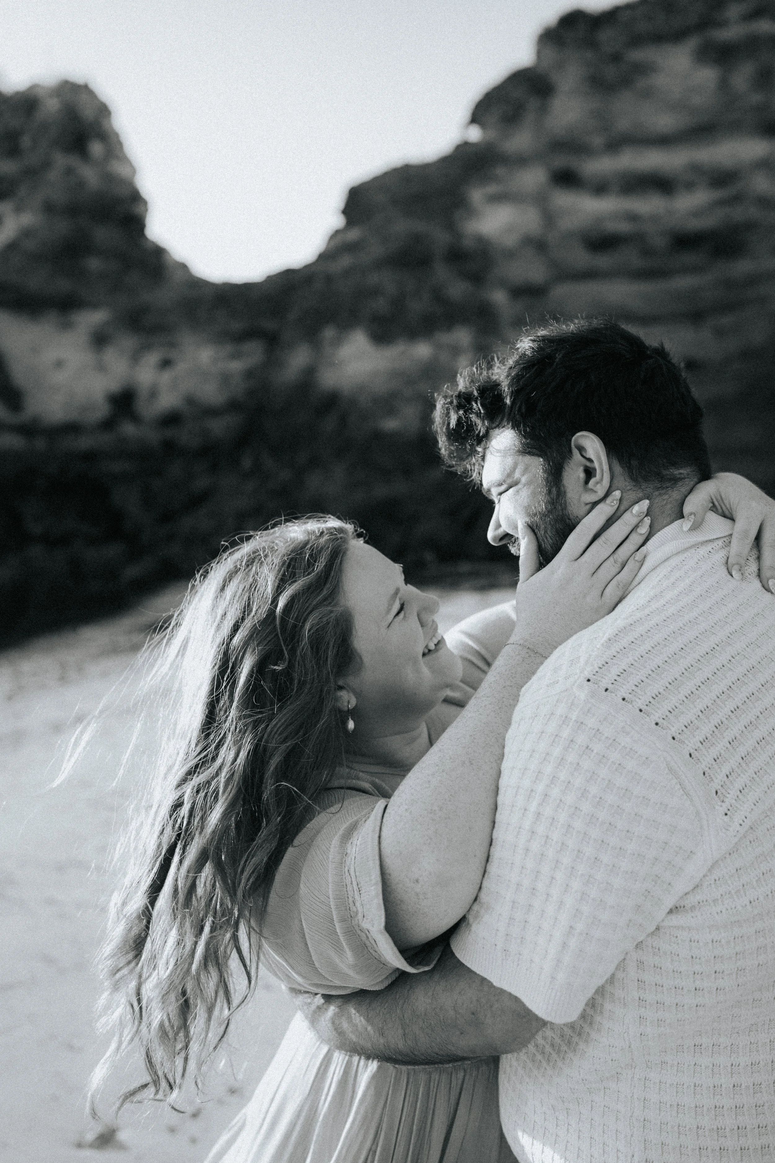 A man and woman share a joyful moment, with the man holding the woman in his arms. They are outdoors against rocky terrain, and both are smiling and looking at each other. The woman has long, curly hair and earrings, and the man has short, dark hair 