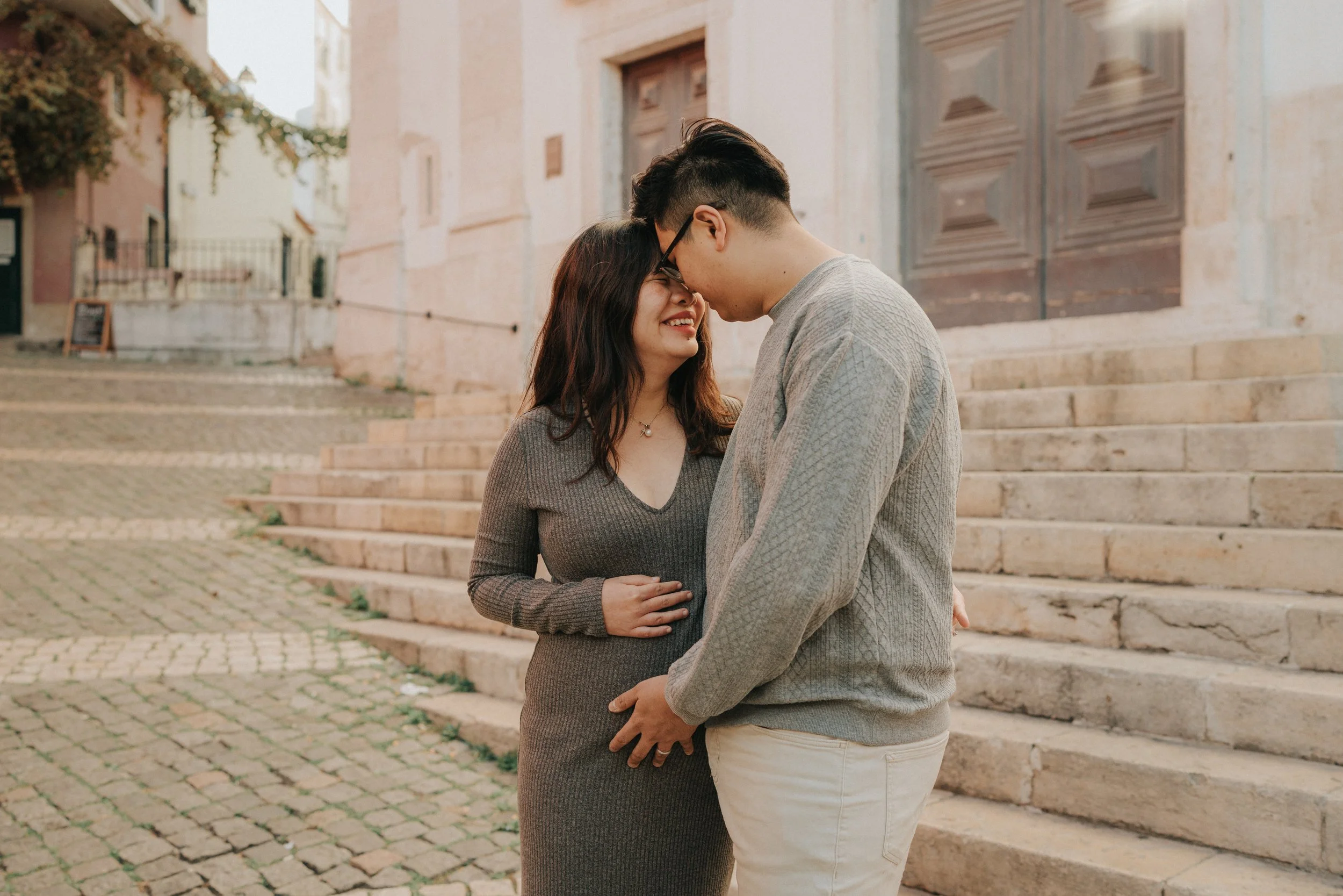 A young couple stands close together on city steps, smiling and touching foreheads as they enjoy a tender moment. The woman has dark hair and glasses, wearing a gray sweater, while the man has dark hair and fair skin, dressed in a light gray sweater 