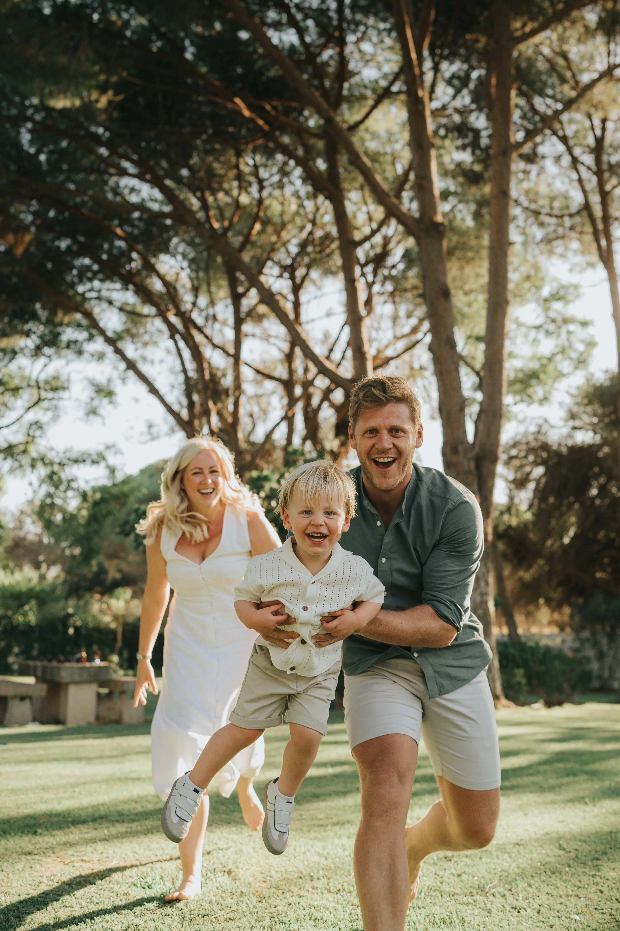 A family of four happily running outdoors in a park on a sunny day, with trees and grass in the background.
