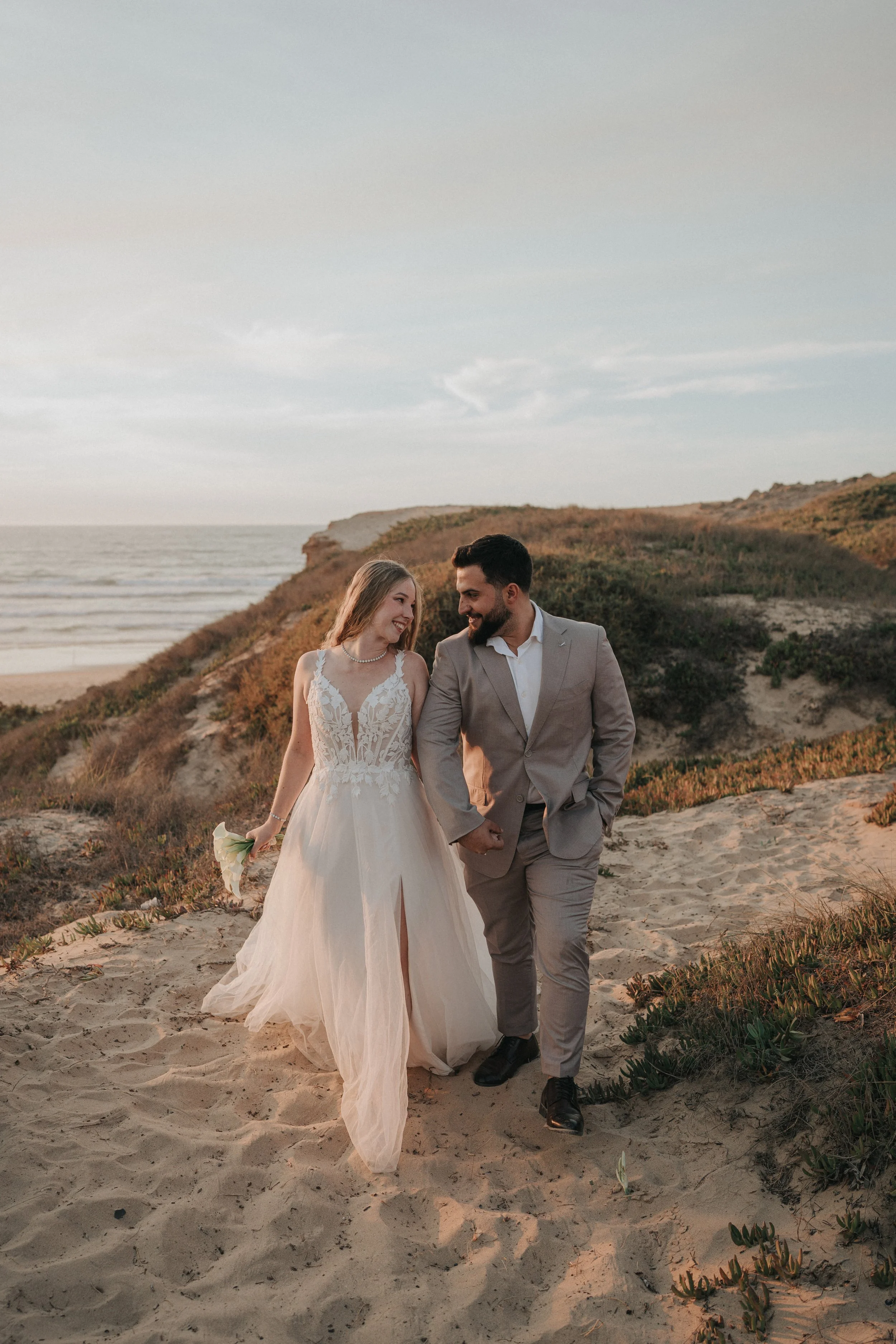 A bride and groom walk hand-in-hand on a sandy beach during sunset, smiling at each other, with dunes and the ocean in the background.
